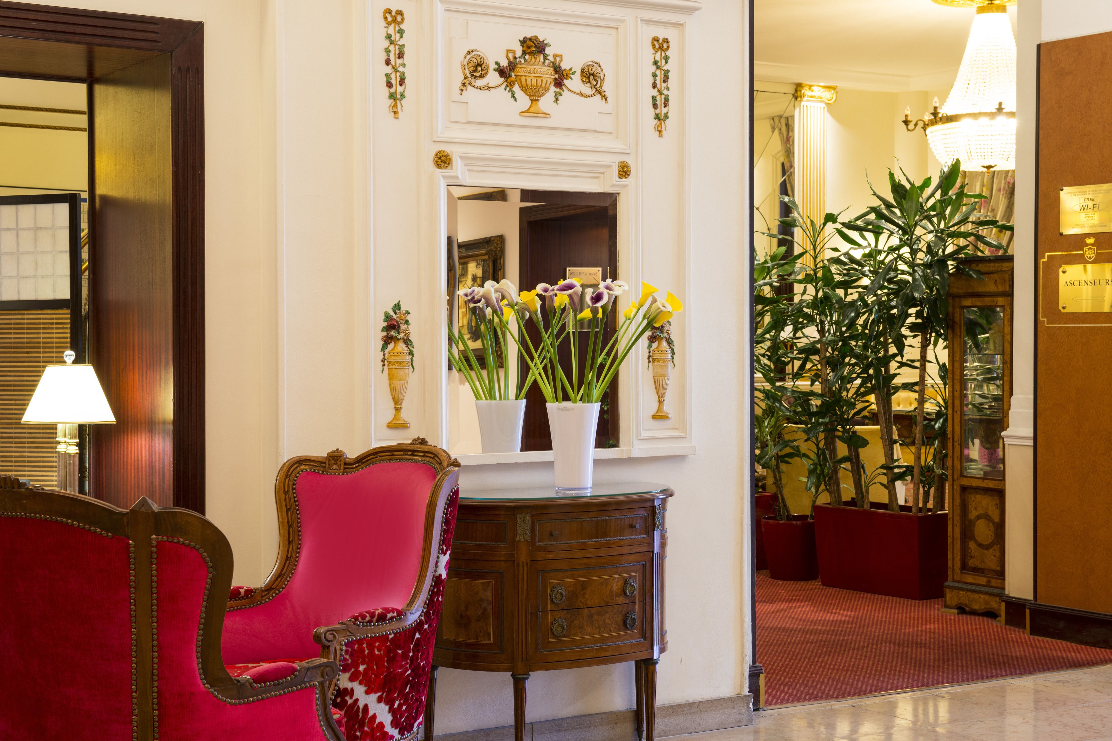 A red upholstered chair, a wooden side table with a vase of flowers, and a large mirror with a decorative frame.