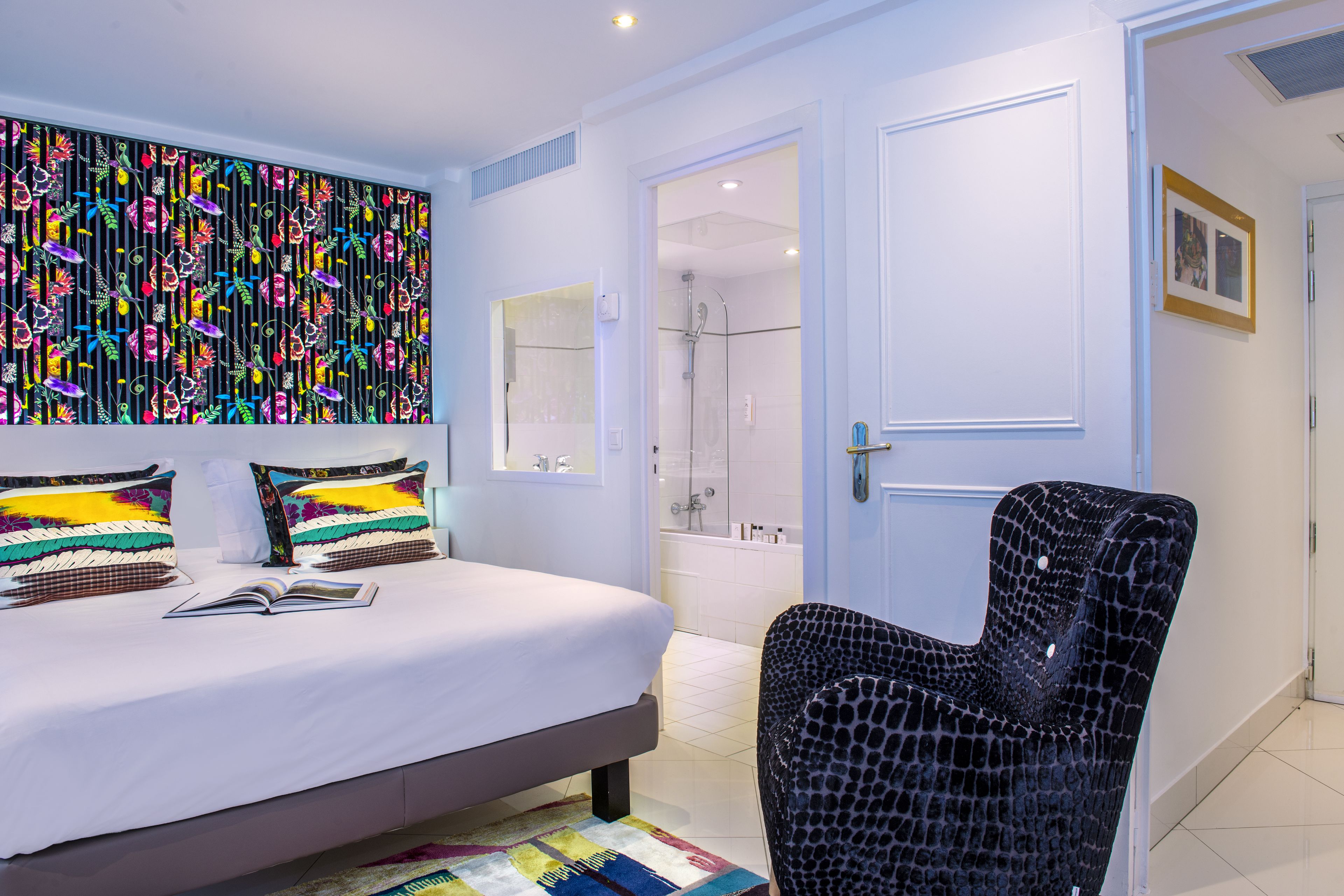 Un dormitorio moderno con cama, almohadas coloridas, una butaca negra con estampado y un baño con ducha y bañera.