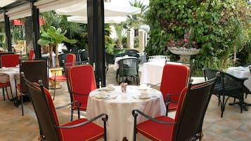 Un espace de restauration en plein air avec des tables rondes et des chaises rouges, entouré de verdure et couvert d’un auvent blanc.