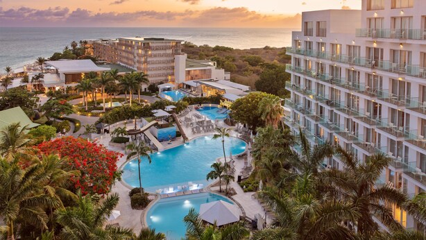 Un resort con varias piscinas, un edificio grande con balcones y vistas al océano al atardecer.
