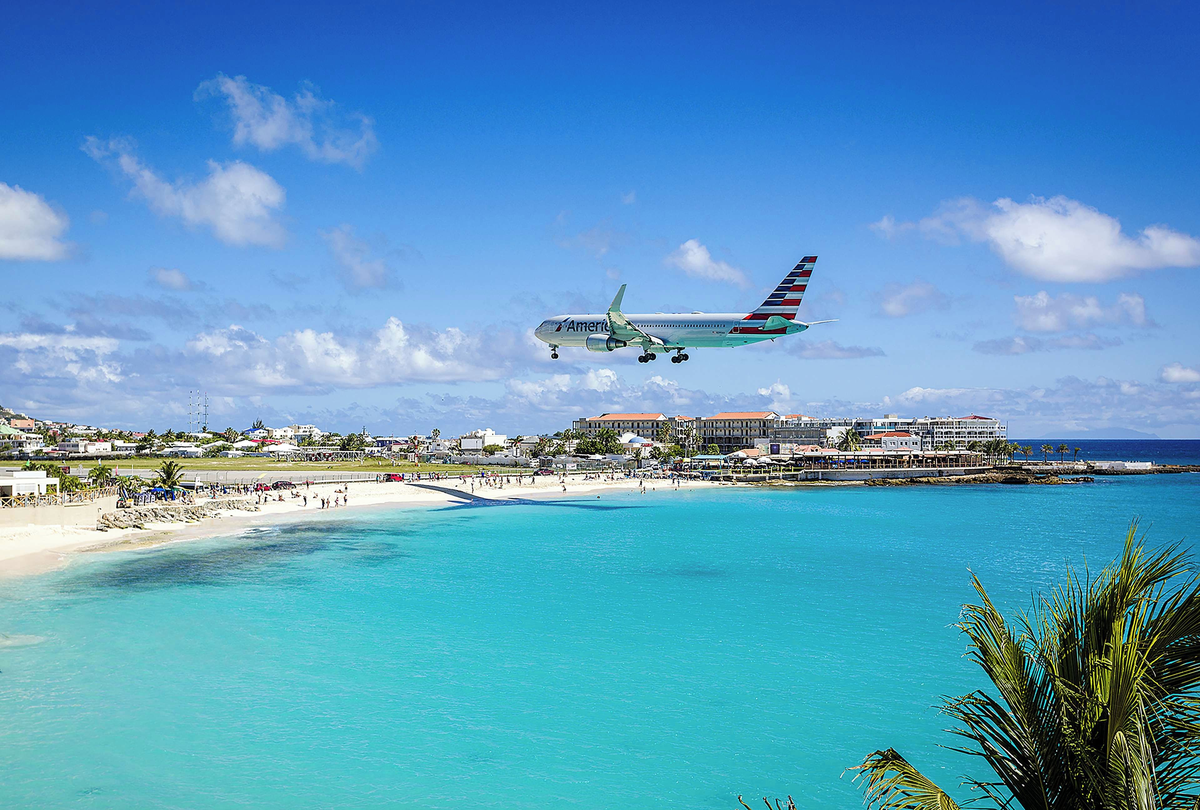 Um avião voando sobre uma praia com água azul cristalina e um resort ao fundo.