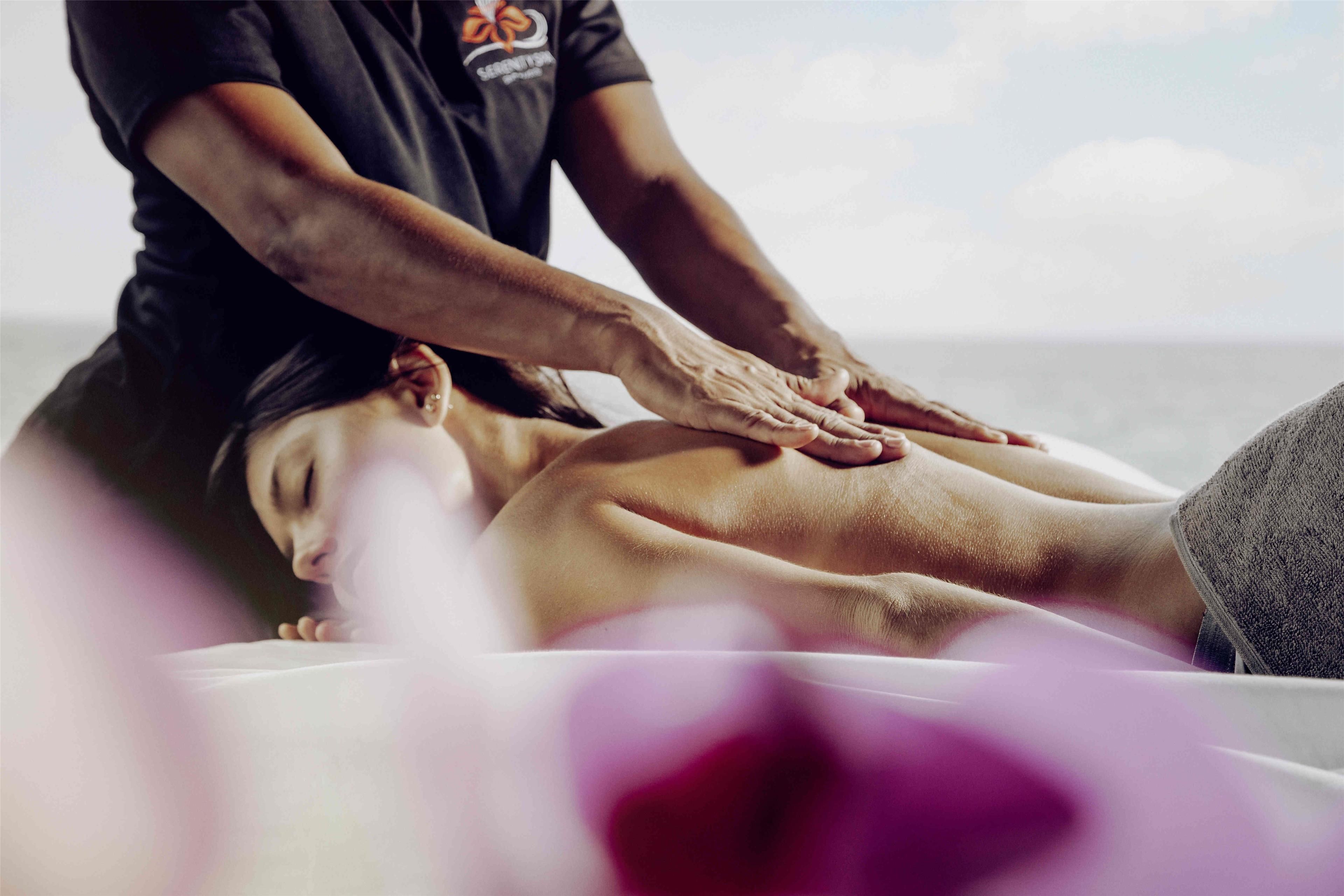 Eine Massageanwendung: Eine Person liegt über einem Behandlungstisch, erhält von einer anderen Person eine Rückenmassage.