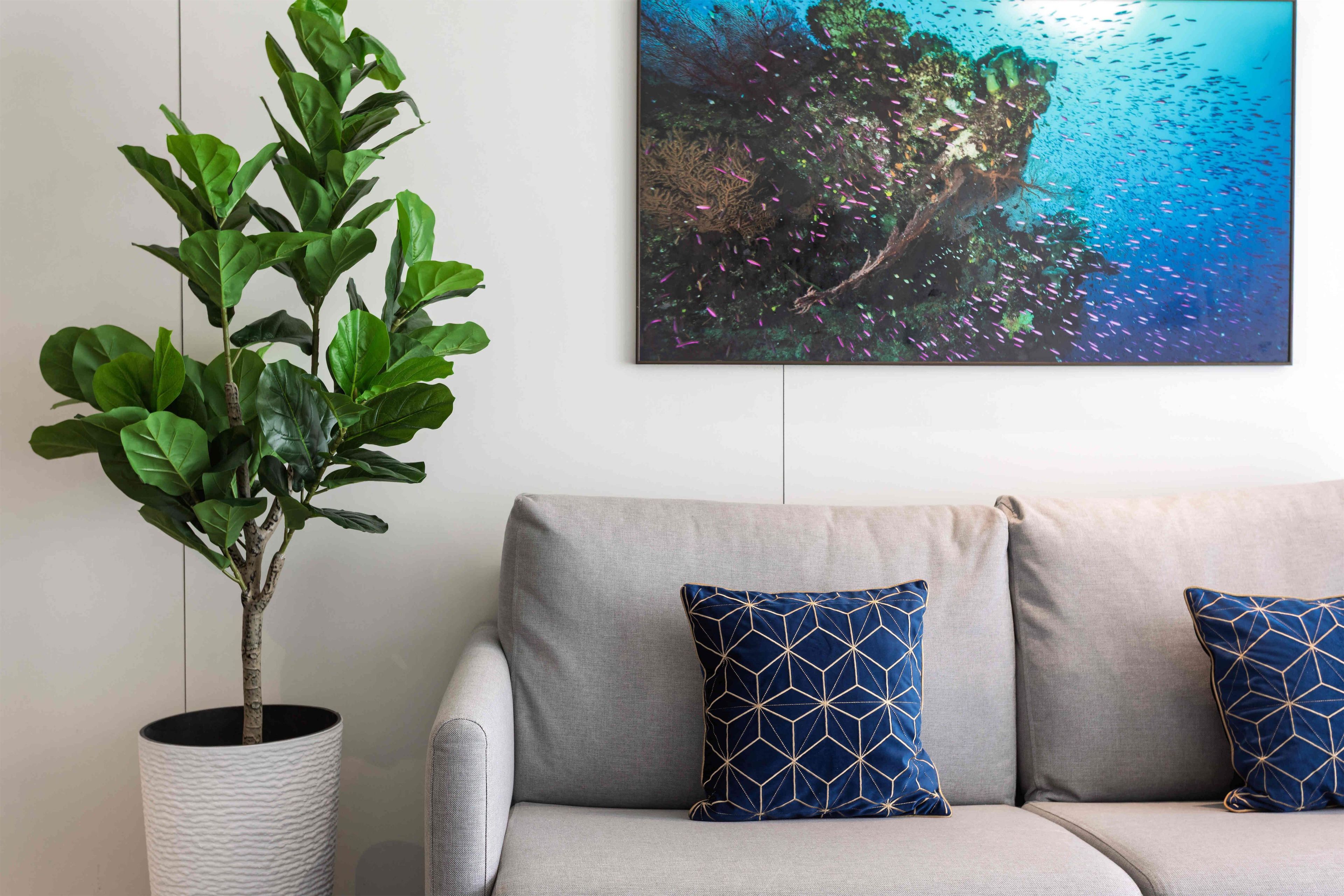 Un sofá gris con dos cojines azules estampados, una planta en maceta y un cuadro colgado en la pared que muestra una escena submarina.