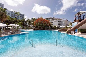 Une piscine entourée de palmiers et bordée de chaises longues et de parasols, avec un bâtiment doté de balcons.