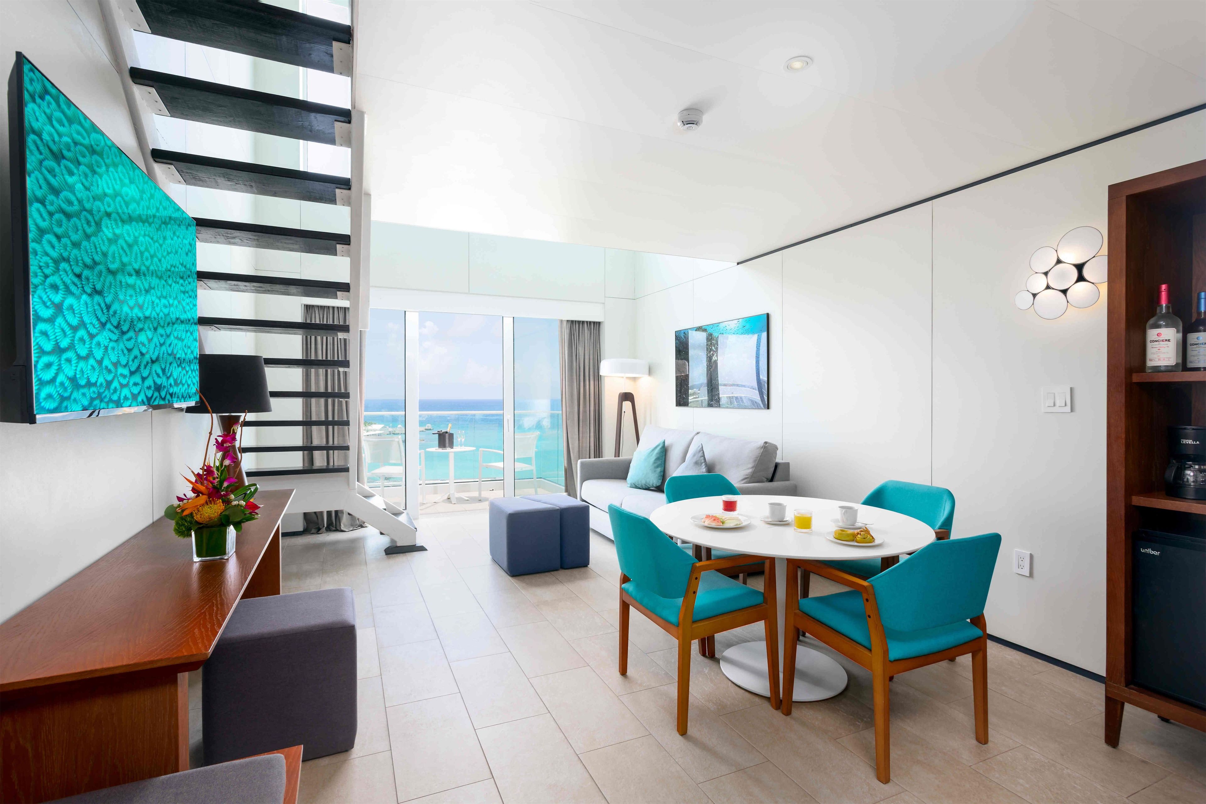 A modern hotel room with a balcony, a dining area with blue chairs, a wooden table, and a kitchen area with a refrigerator.