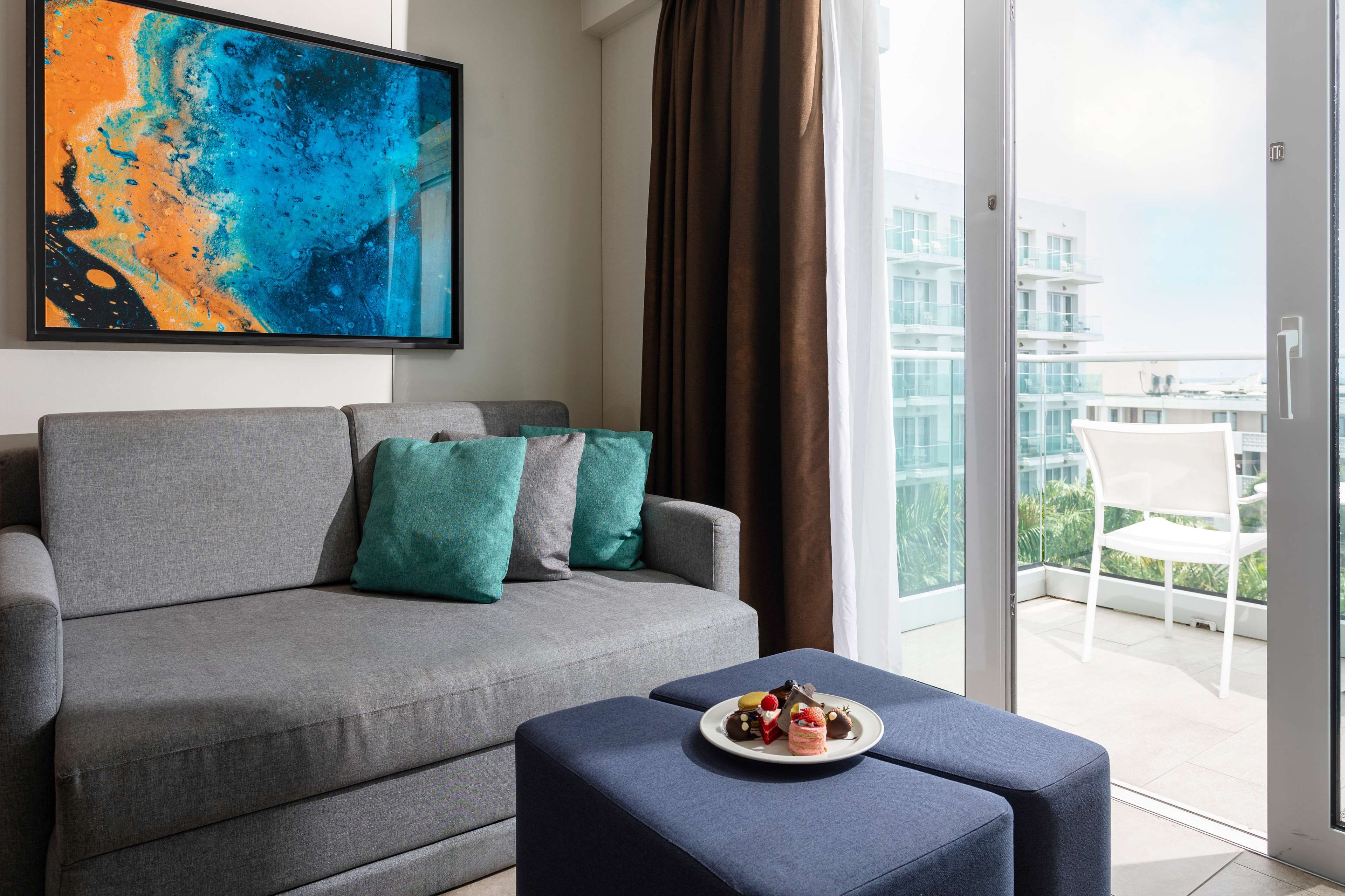 A modern living room with a grey sofa, teal cushions, a coffee table with a plate of pastries, and a large abstract painting.