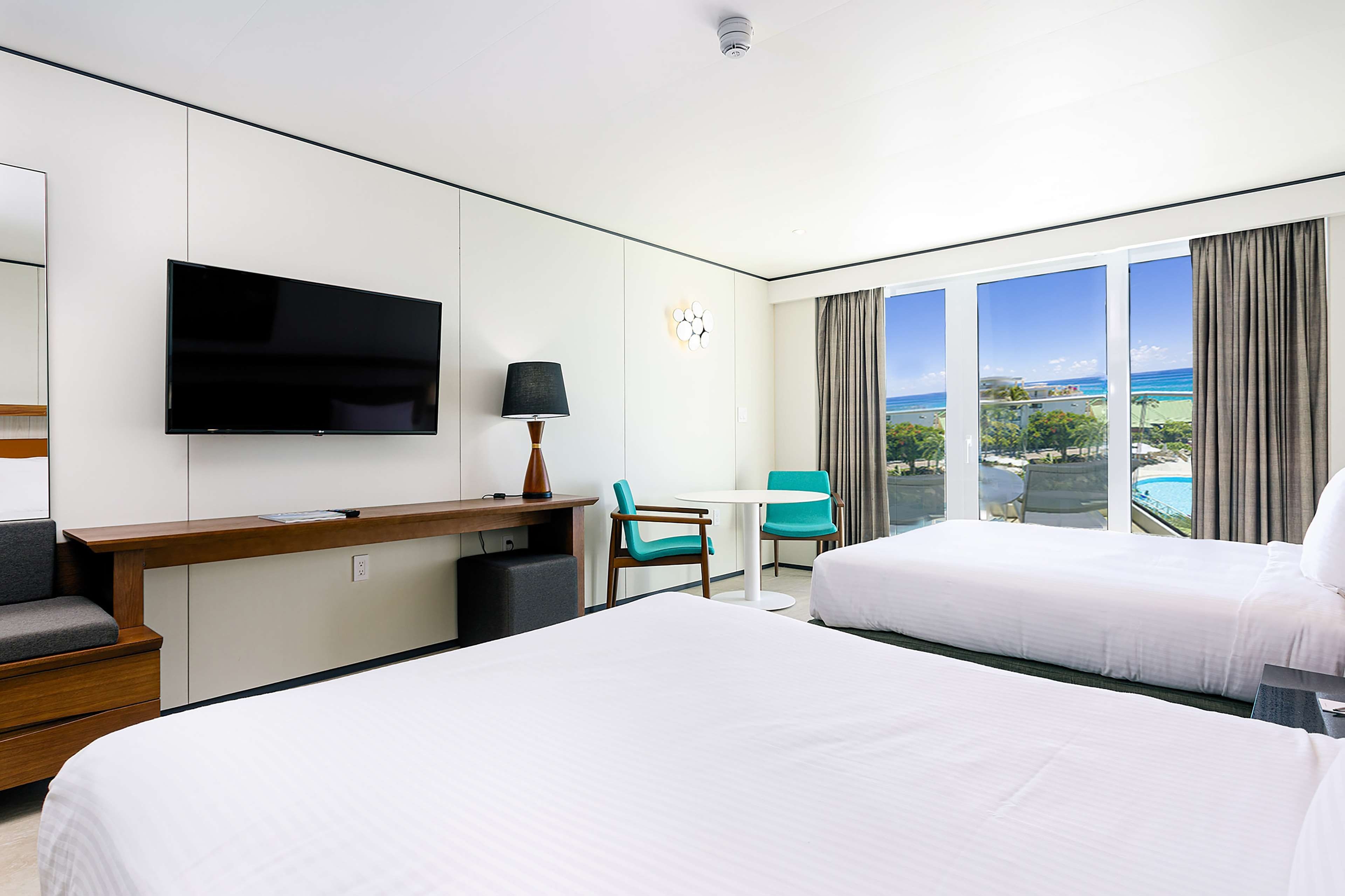 A hotel room with two beds, a desk with a flat-screen TV, a chair, and a view of the ocean.