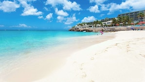 Una playa con aguas turquesas cristalinas, una orilla de arena y un hotel con varios pisos y palmeras.