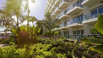 A modern hotel with a lush garden, balconies with glass railings, and a clear view of the sky.