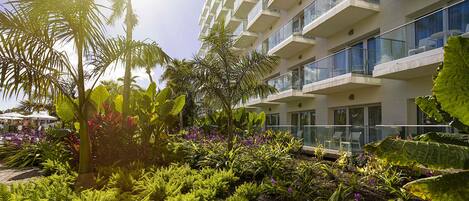 A modern hotel with a lush garden, balconies with glass railings, and a clear view of the sky.