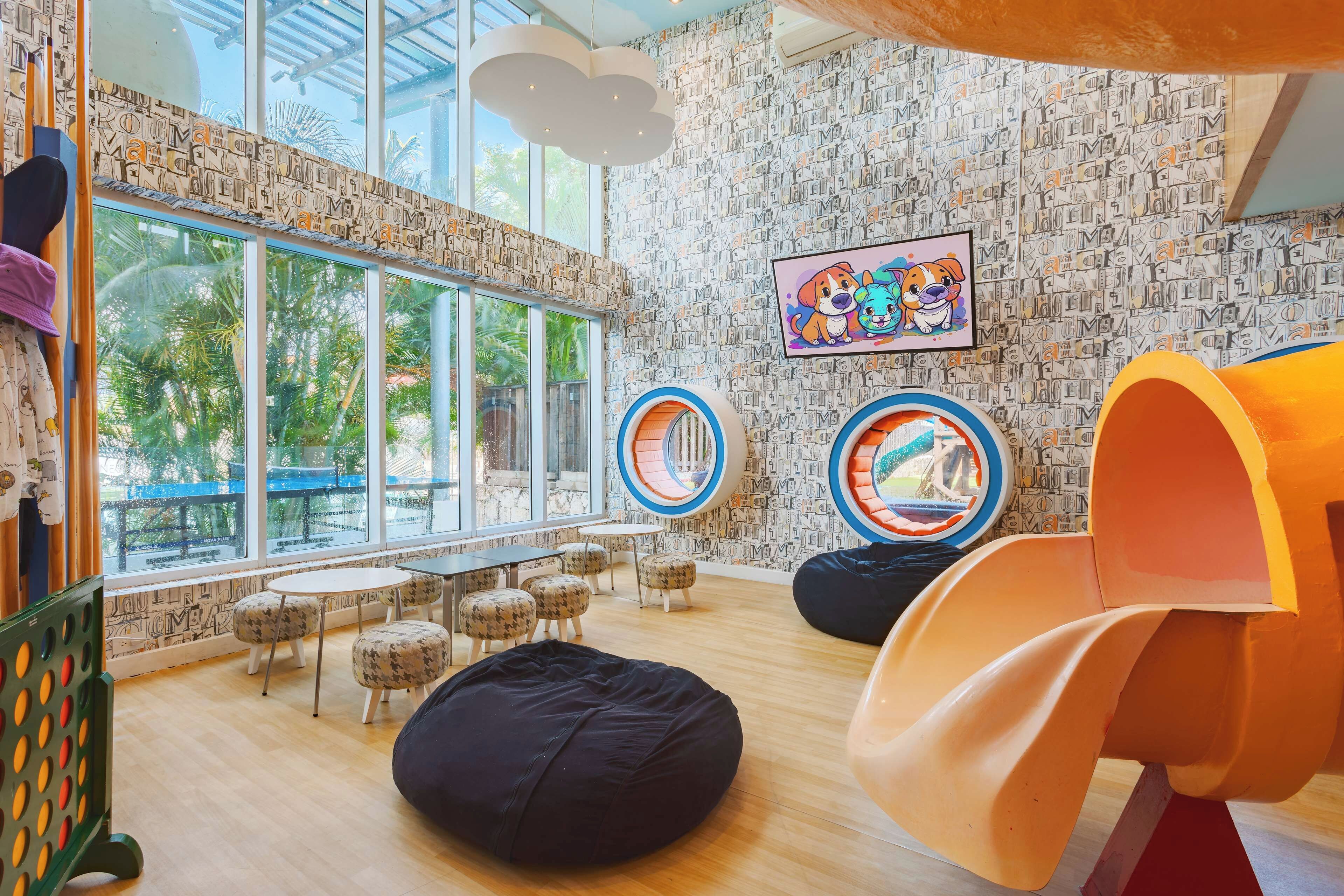 A children's play area with bean bags, circular play structures, and a large window with a view of palm trees.