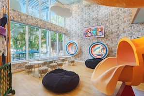 Zona de juegos infantiles con sillones de espuma, estructuras circulares y un amplio ventanal con vistas a palmeras.