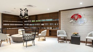 Zona de recepción de un spa con un mostrador de madera, sillas blancas y una pared con un logotipo y estantes de productos.