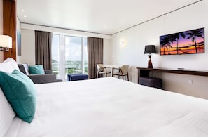 Ein modernes Hotelzimmer mit einem großen Bett, einem Schreibtisch, einem Sessel und Blick auf die Stadt durch große Fenster.