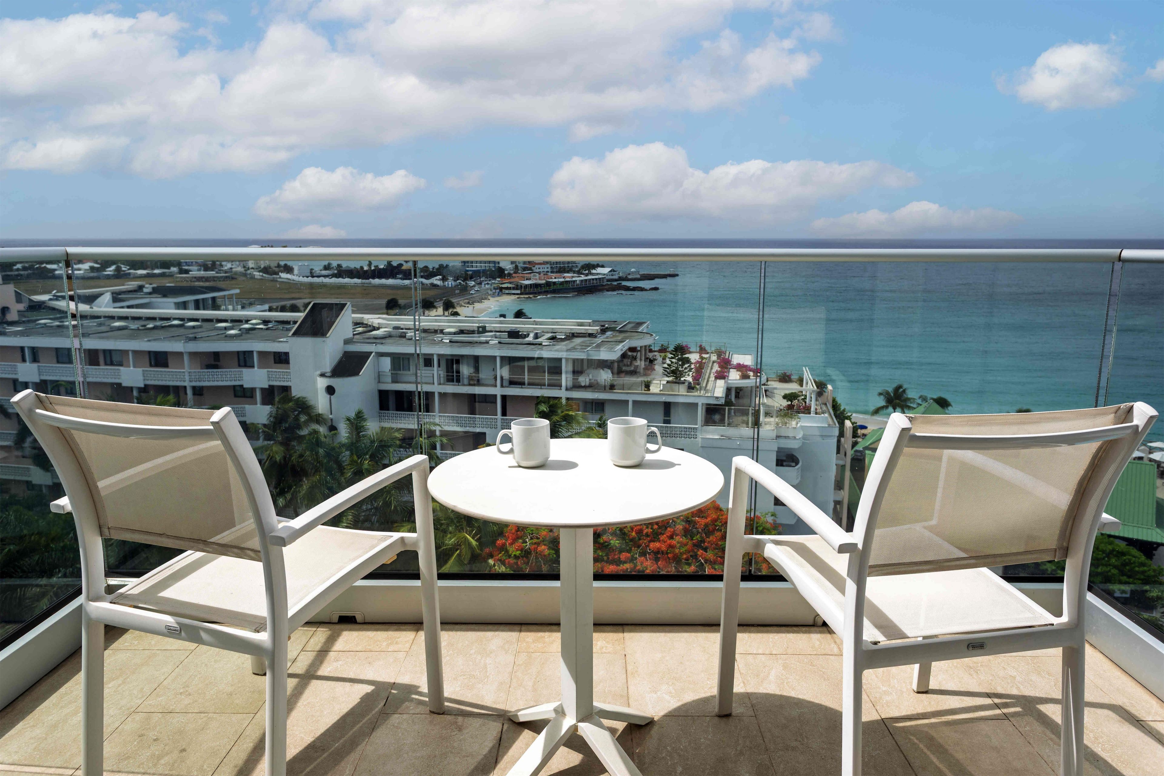 A balcony with two white chairs and a small table, overlooking a coastal area with buildings and a beach.