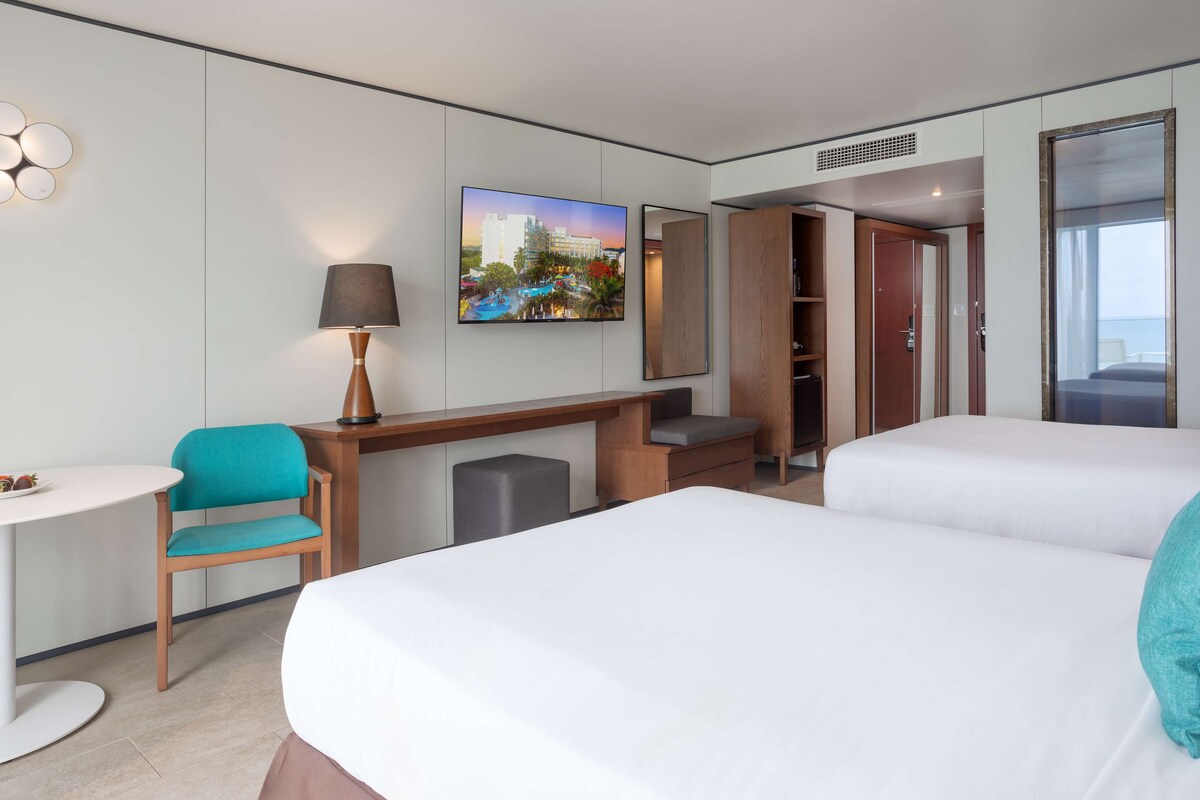 Ein Hotelzimmer mit zwei Betten, einem Schreibtisch, einem Stuhl, einer Lampe, einem Fernseher und einem Kleiderschrank.