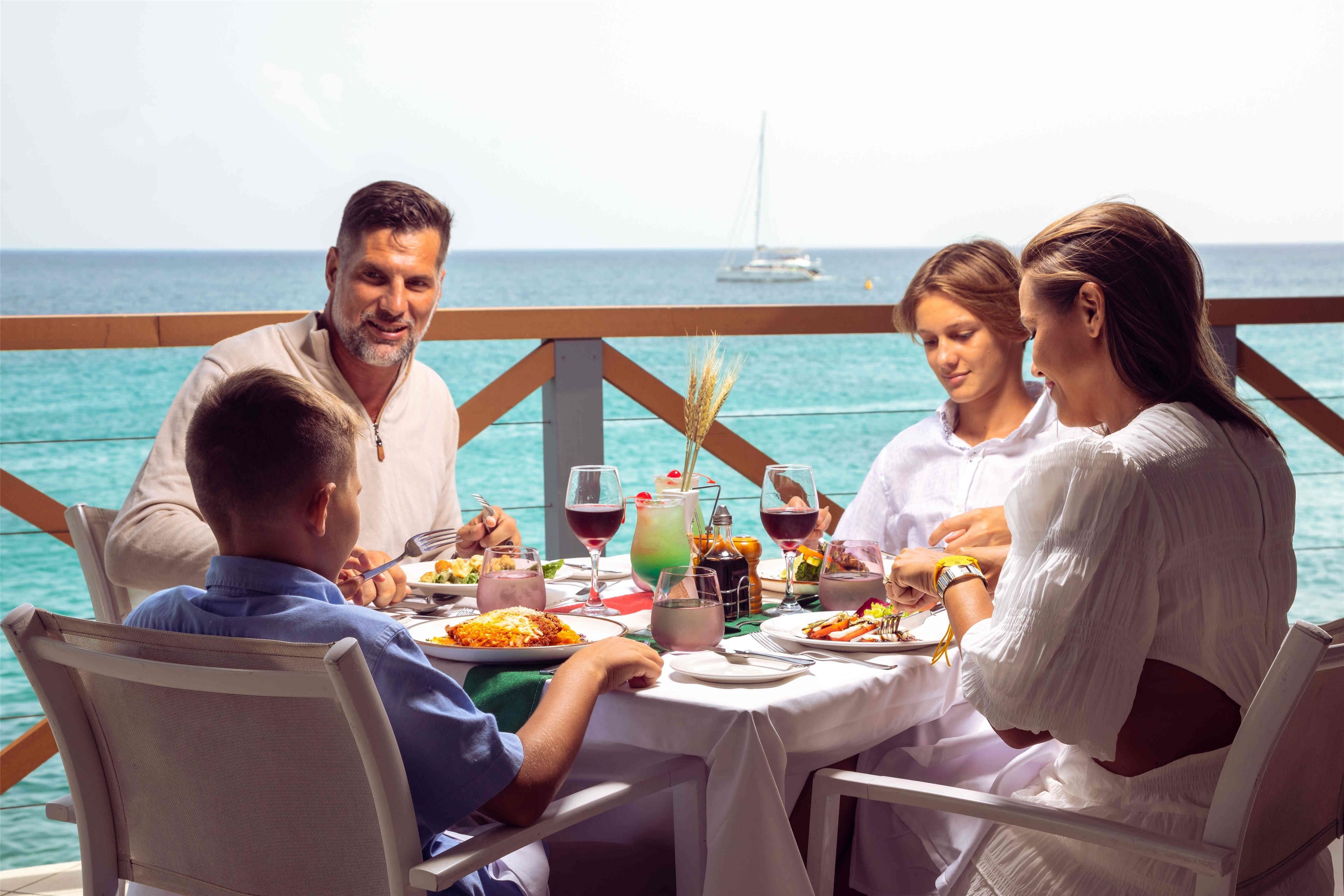 Un grupo cenando en una mesa con vista al mar y un velero al fondo.
