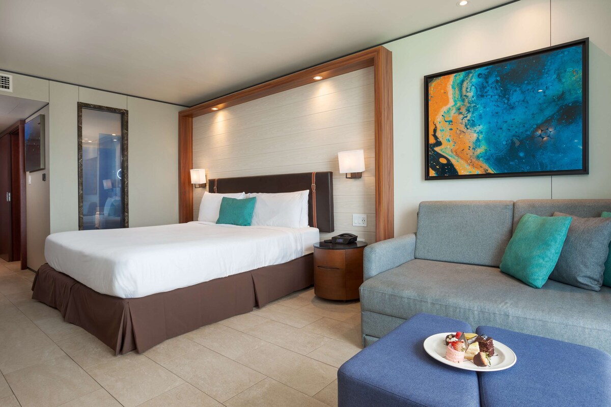 Ein Hotelzimmer mit einem großen Bett, einem Sofa, einem Nachttisch mit Lampe und einer Platte mit Desserts auf dem Sofa.