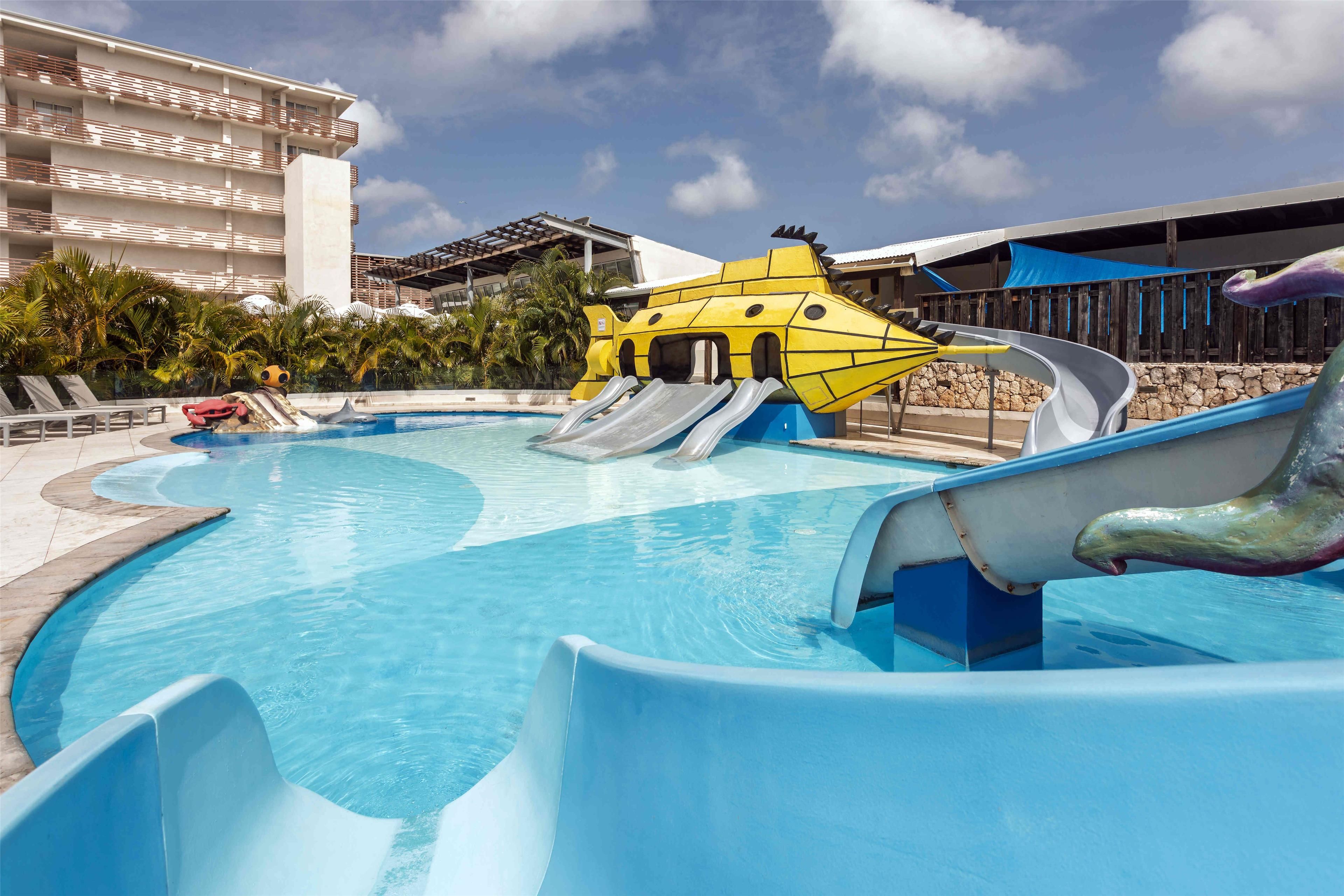 A swimming pool with slides and a water play area, surrounded by palm trees and a building with balconies.