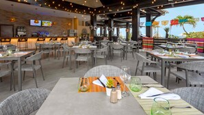 Un restaurante con terraza, mesas dispuestas con cristalería y cubiertos, y una zona de bar con televisor y dispensadores de bebidas.
