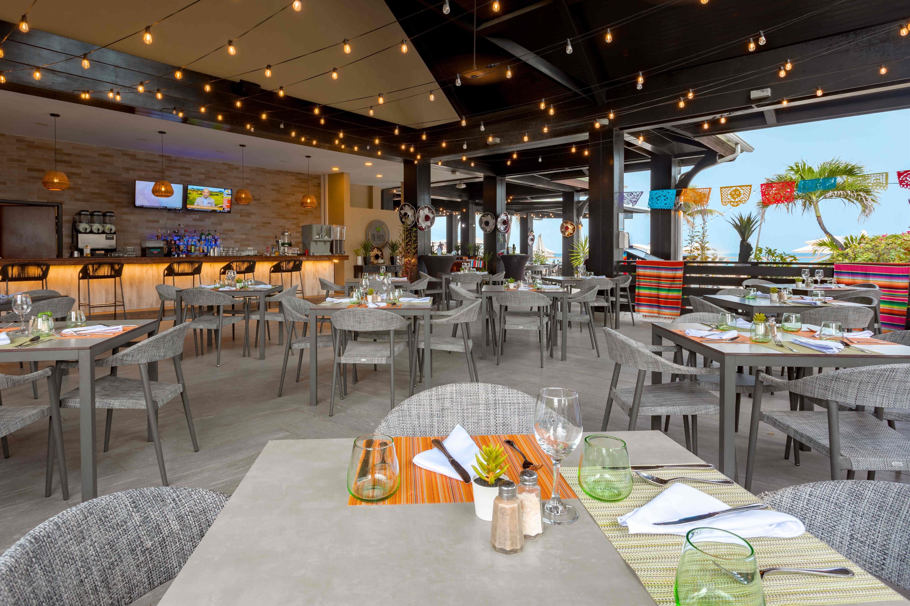 A restaurant with outdoor seating, tables set with glassware and cutlery, and a bar area with a television and beverage dispensers.