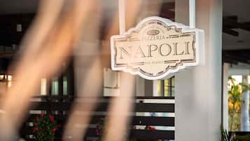 Placa para 'NAPOLI PIZZERIA' com design clássico, localizada em um edifício moderno com colunas estruturais aparentes e um banco ao fundo.