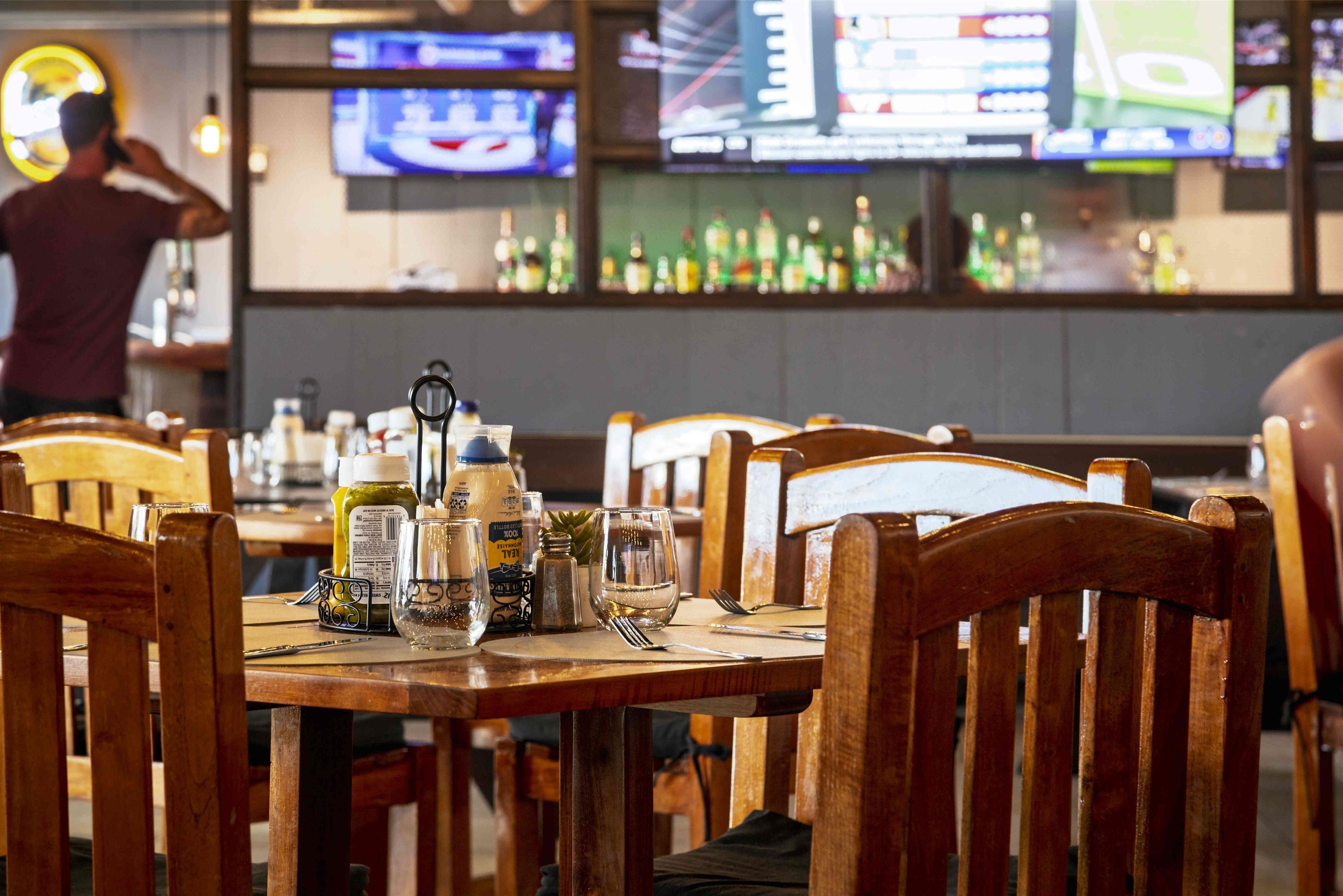 A restaurant with wooden tables and chairs, a bar area with bottles, and multiple screens displaying sports content.