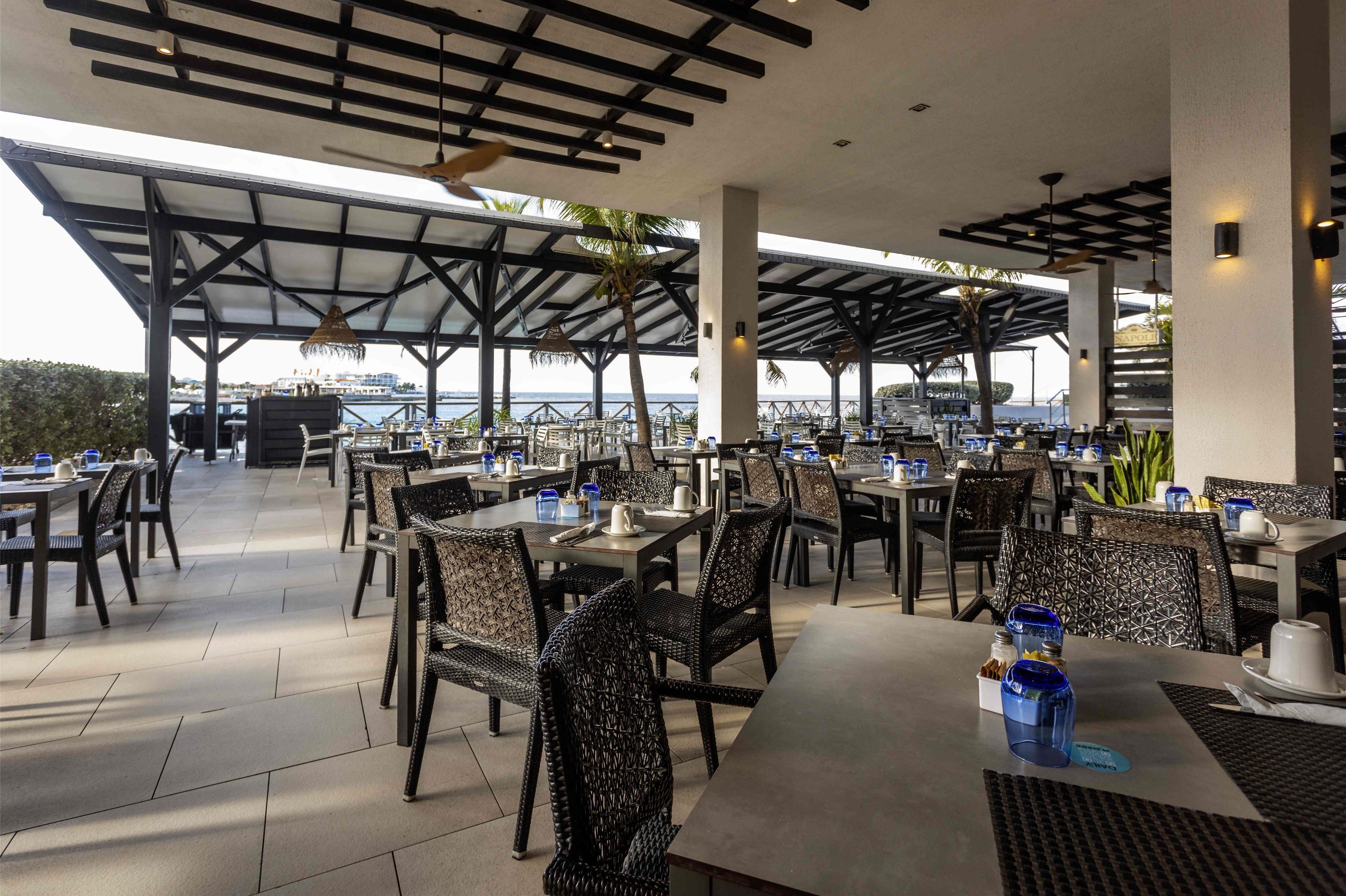 Un espace extérieur aménagé pour les repas, avec des tables et des chaises, un toit abrité et une vue sur la mer.