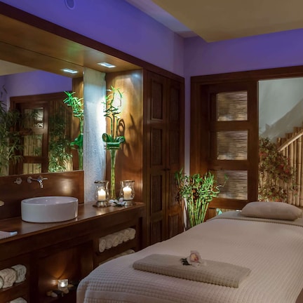 A well-lit bathroom with a large mirror, a sink, and a bathtub. There are multiple plants and candles on the vanity.