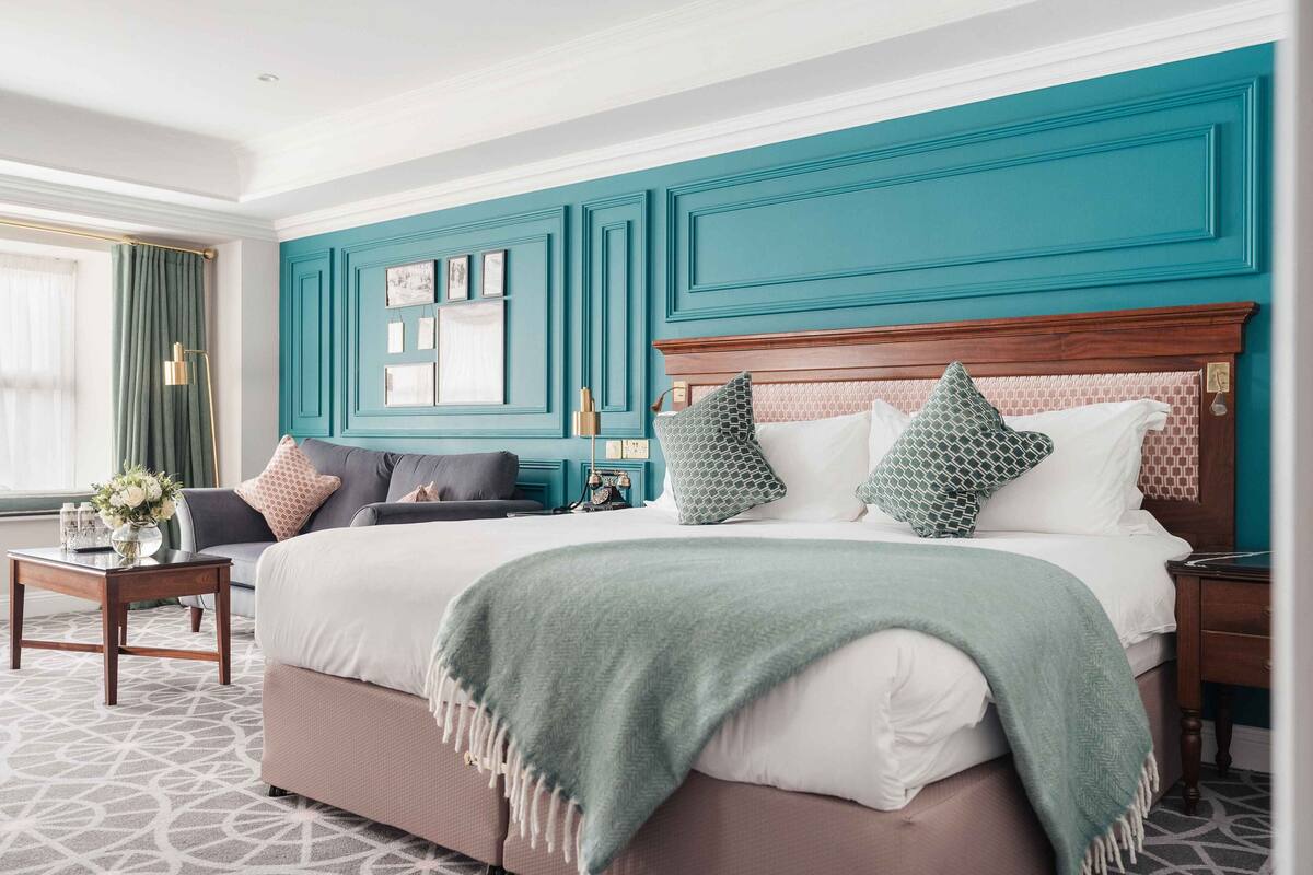 A hotel room with a large bed, a bedside table with a vase of flowers, a sofa with cushions, and a view of the city.