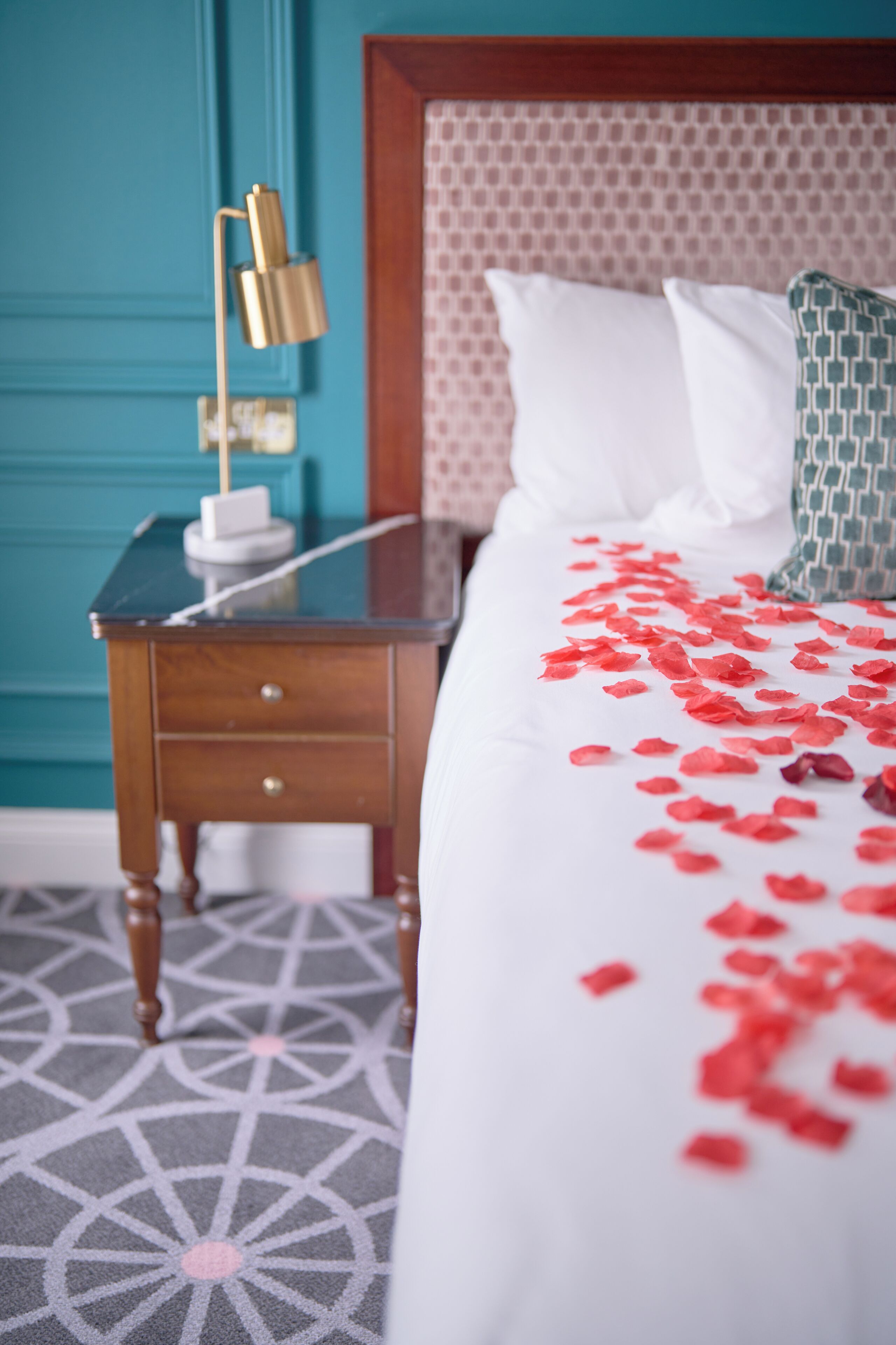 A neatly made bed with white bedding and red rose petals scattered on it, a bedside table with a lamp, and a patterned carpet.