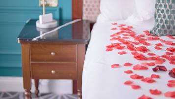 A neatly made bed with white bedding and red rose petals scattered on it, a bedside table with a lamp, and a patterned carpet.