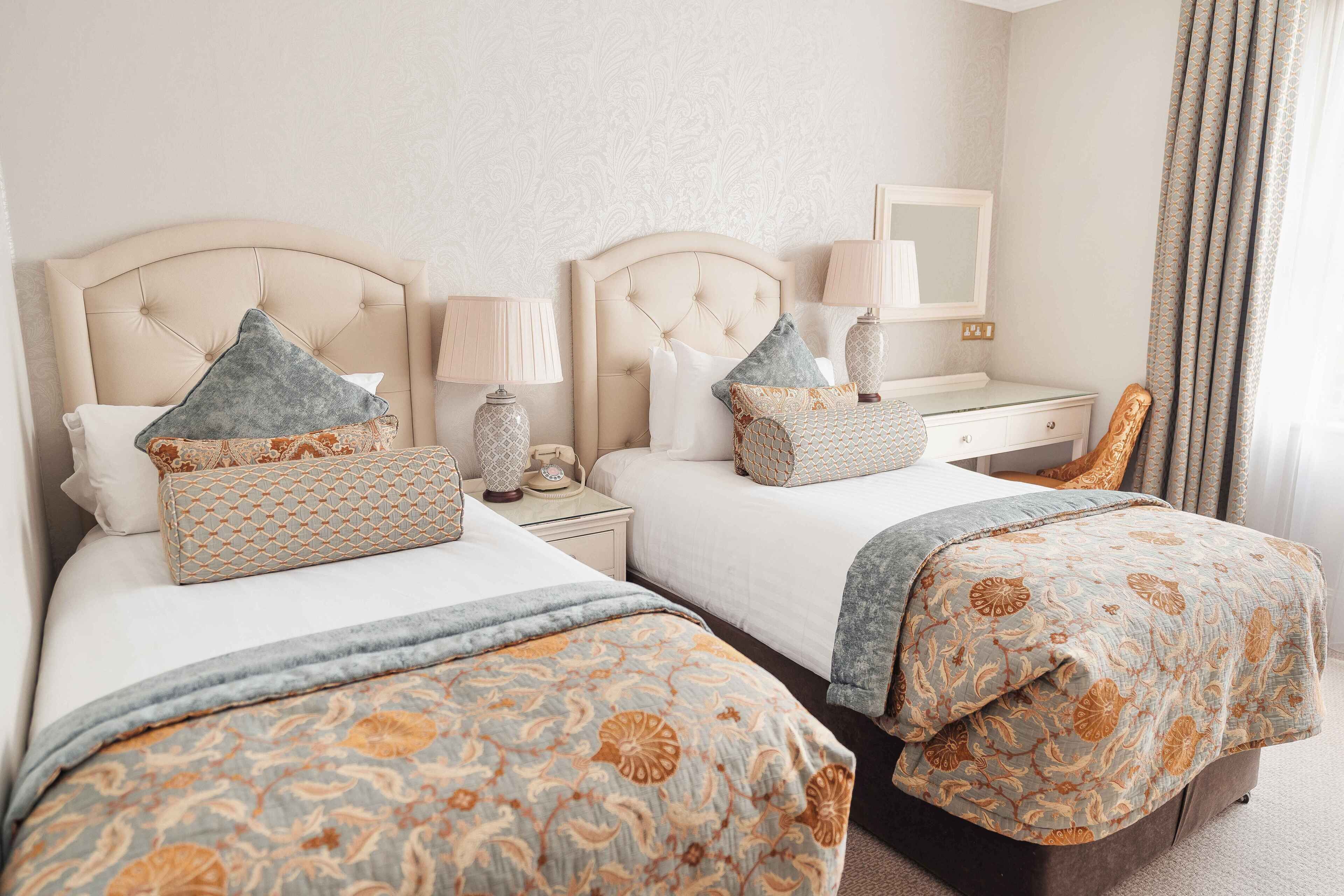 A hotel room with two beds, each with a headboard, bedside tables with lamps, and a mirror above the dresser.