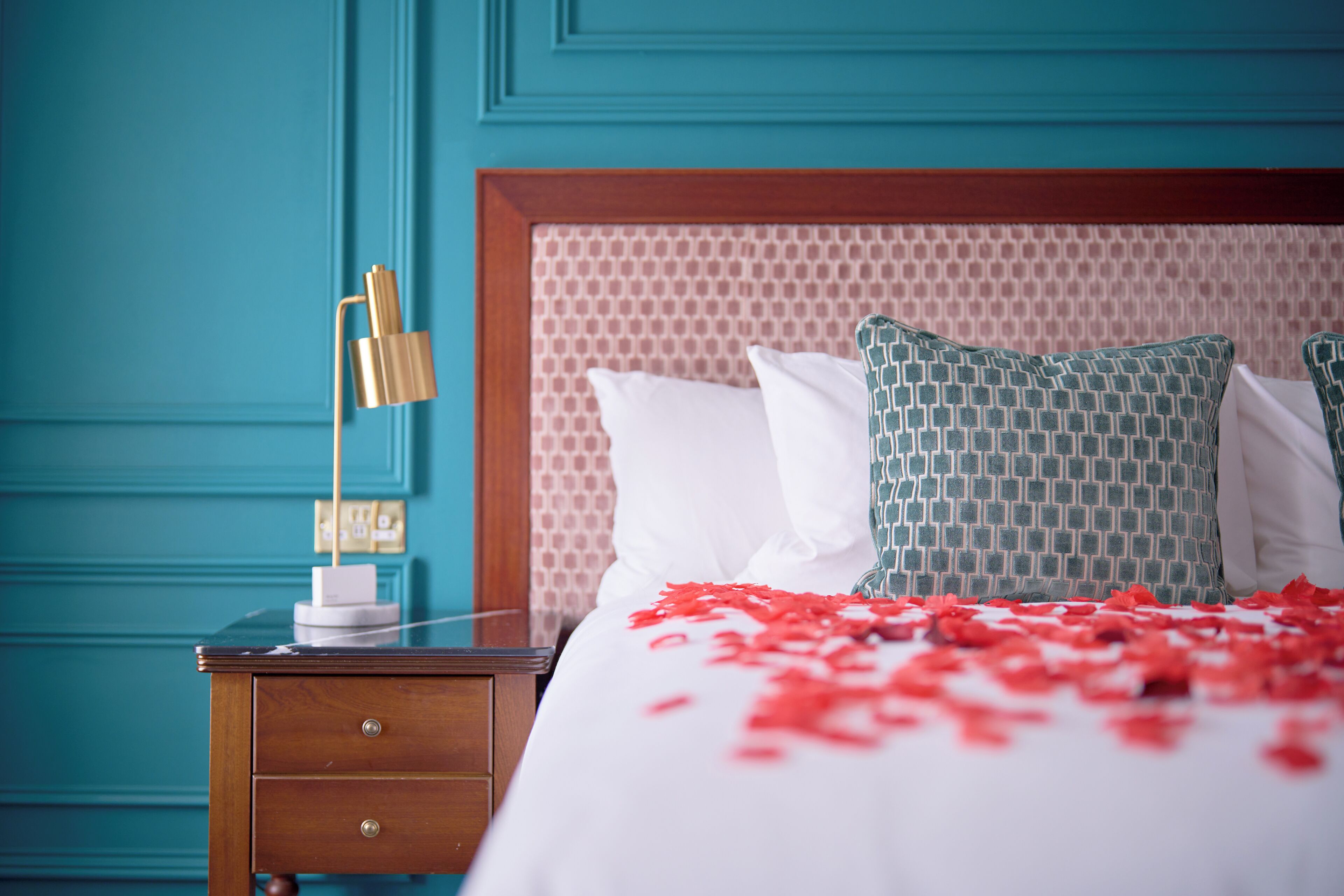 A neatly made bed with white and patterned pillows, a wooden nightstand with a lamp, and a teal wall.