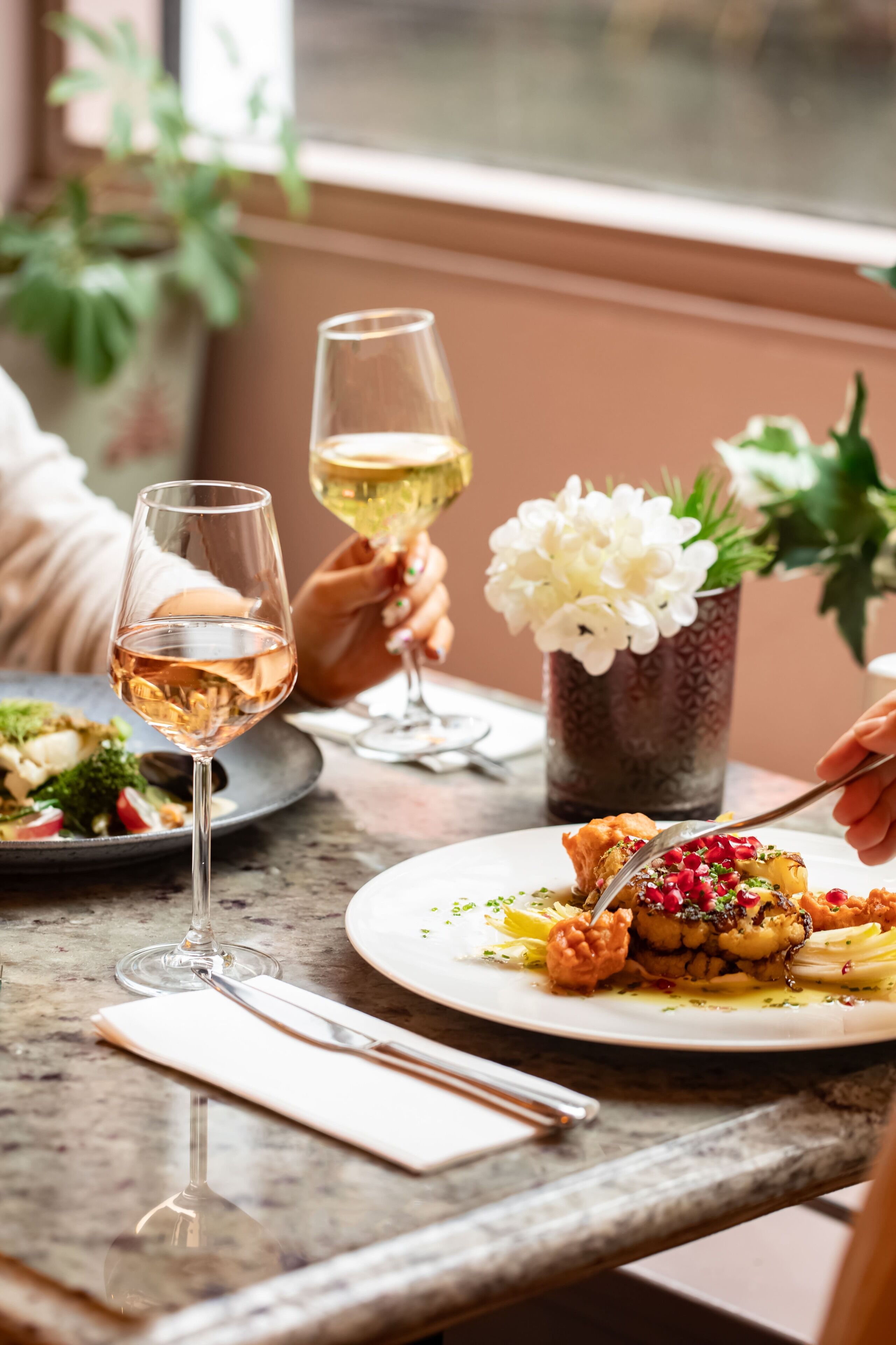 Une table à manger avec deux verres à vin, une assiette de nourriture et un vase rempli de fleurs.