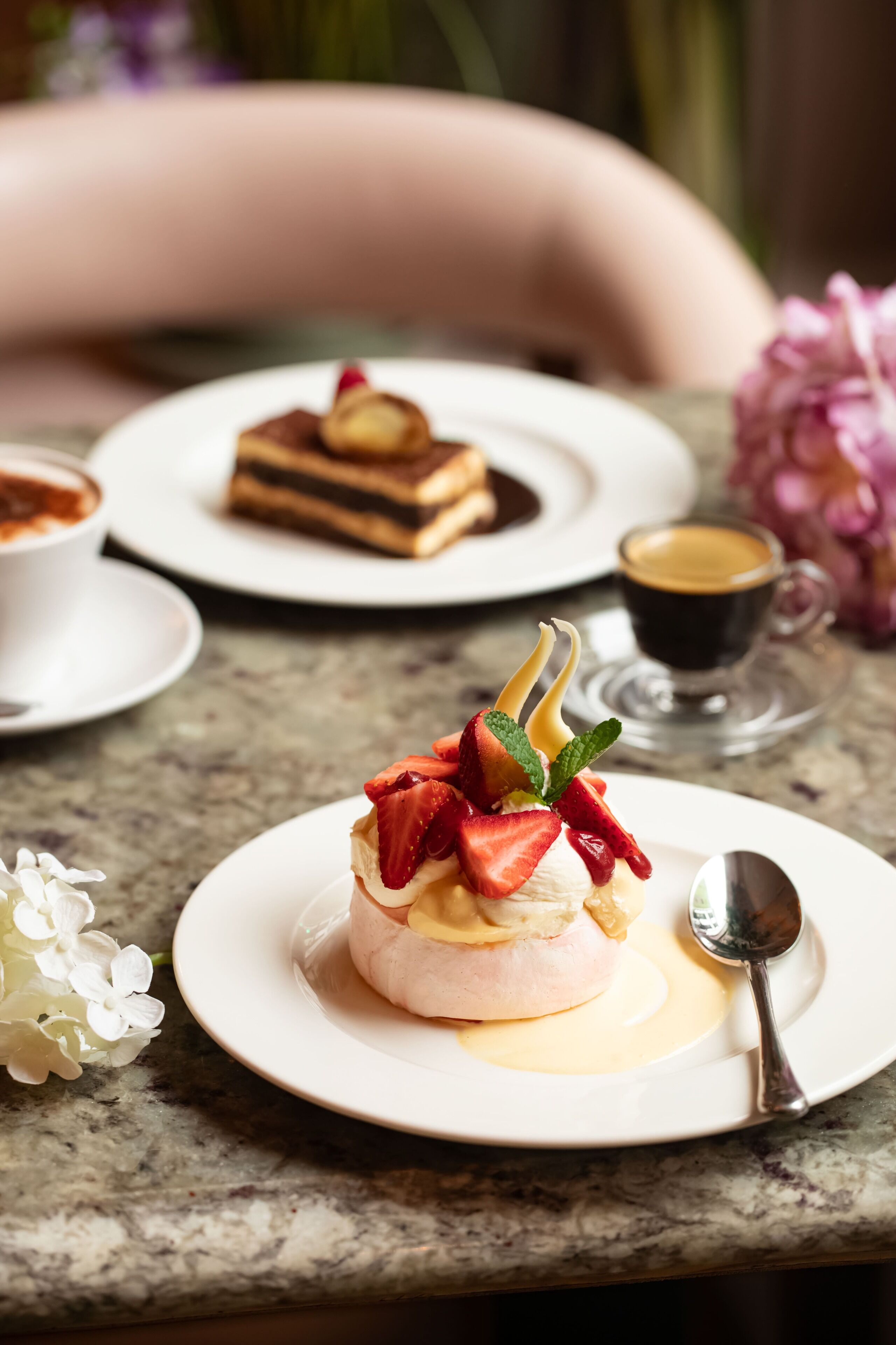 A dessert plate with a meringue topped with strawberries and mint, a coffee cup, and a slice of cake on a marble table.