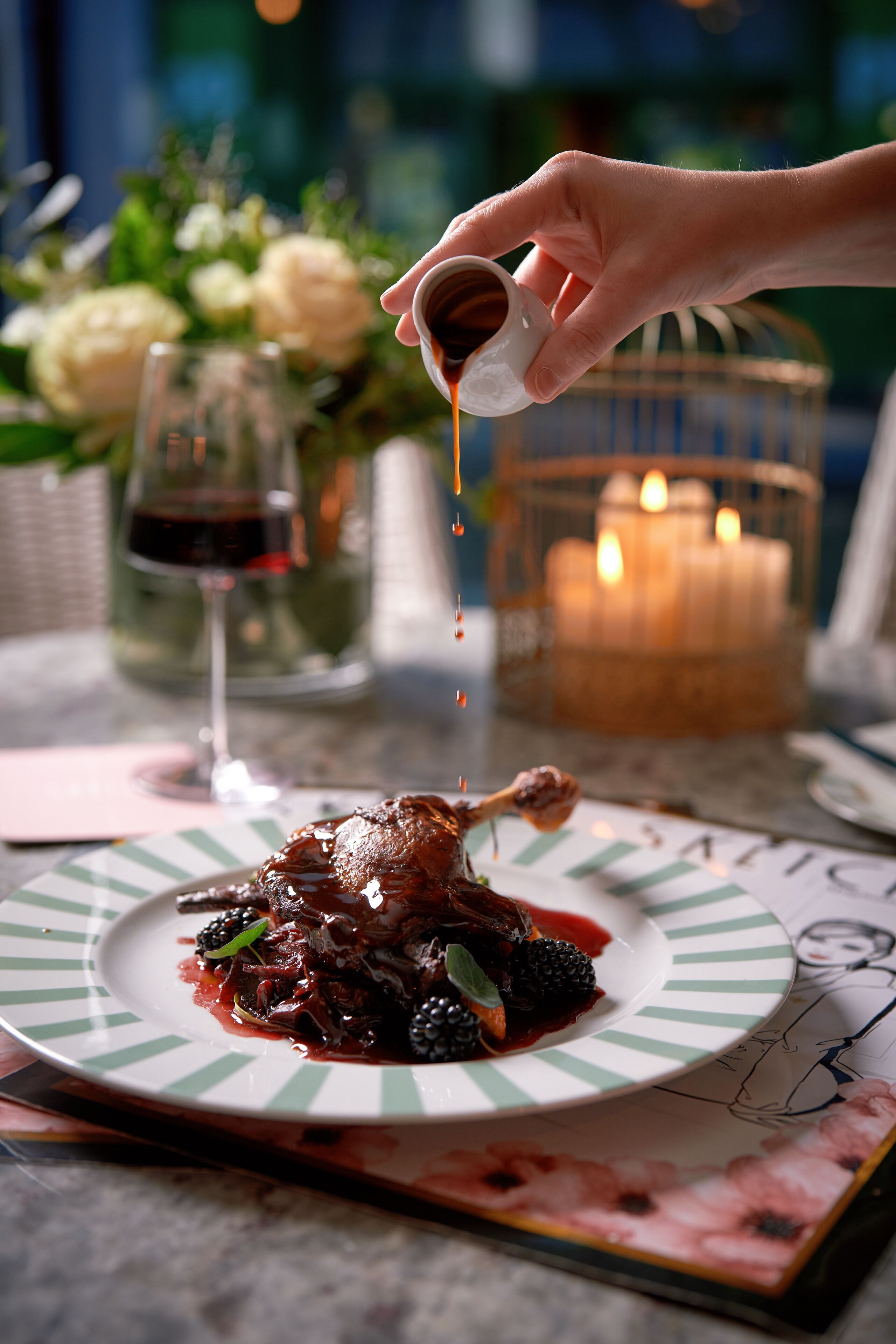 A hand pouring a dark liquid onto a plate with a striped rim, a candle, and a glass of red wine in the background.