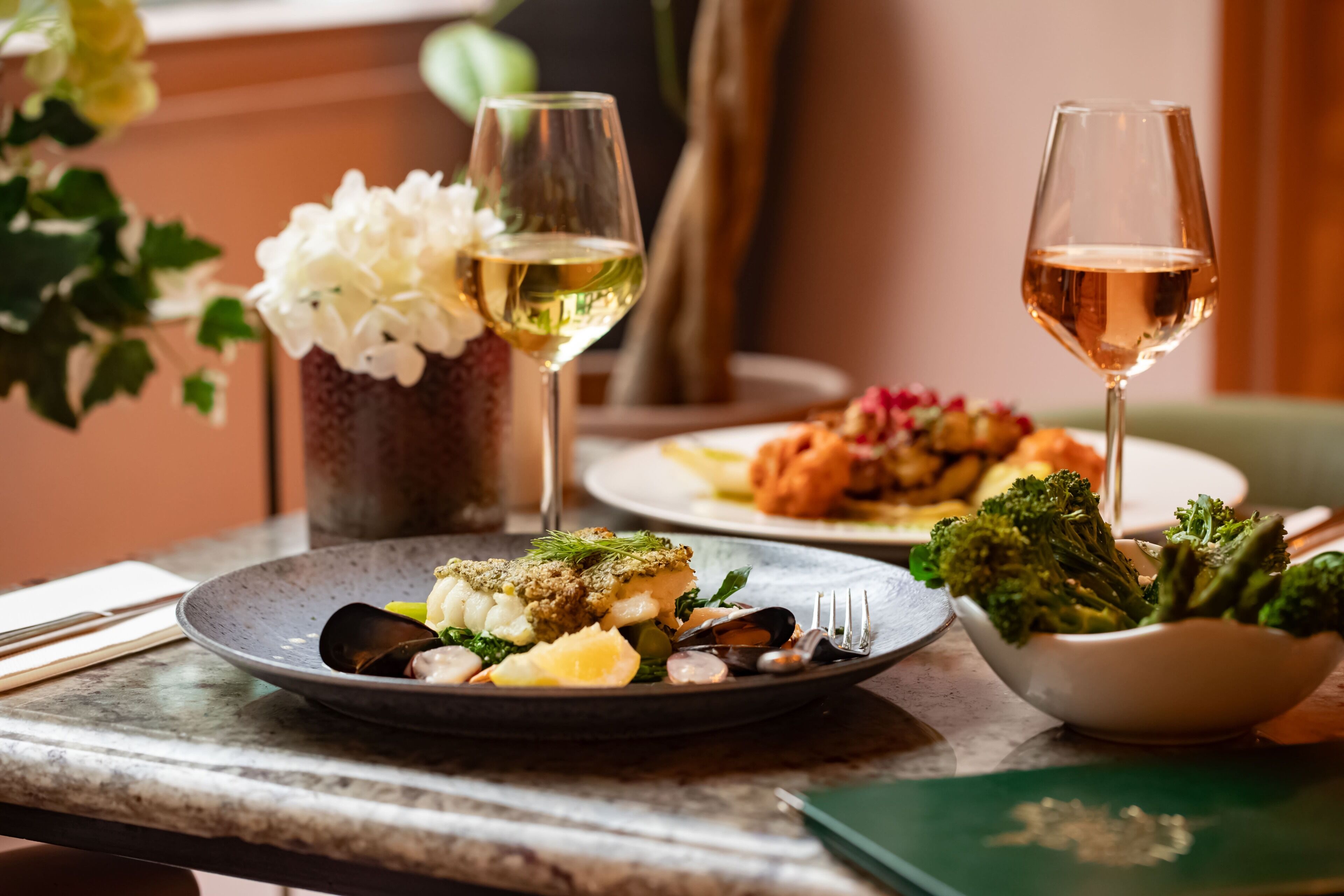Una mesa de comedor con platos de comida, dos copas de vino blanco y un tazón de brócoli.