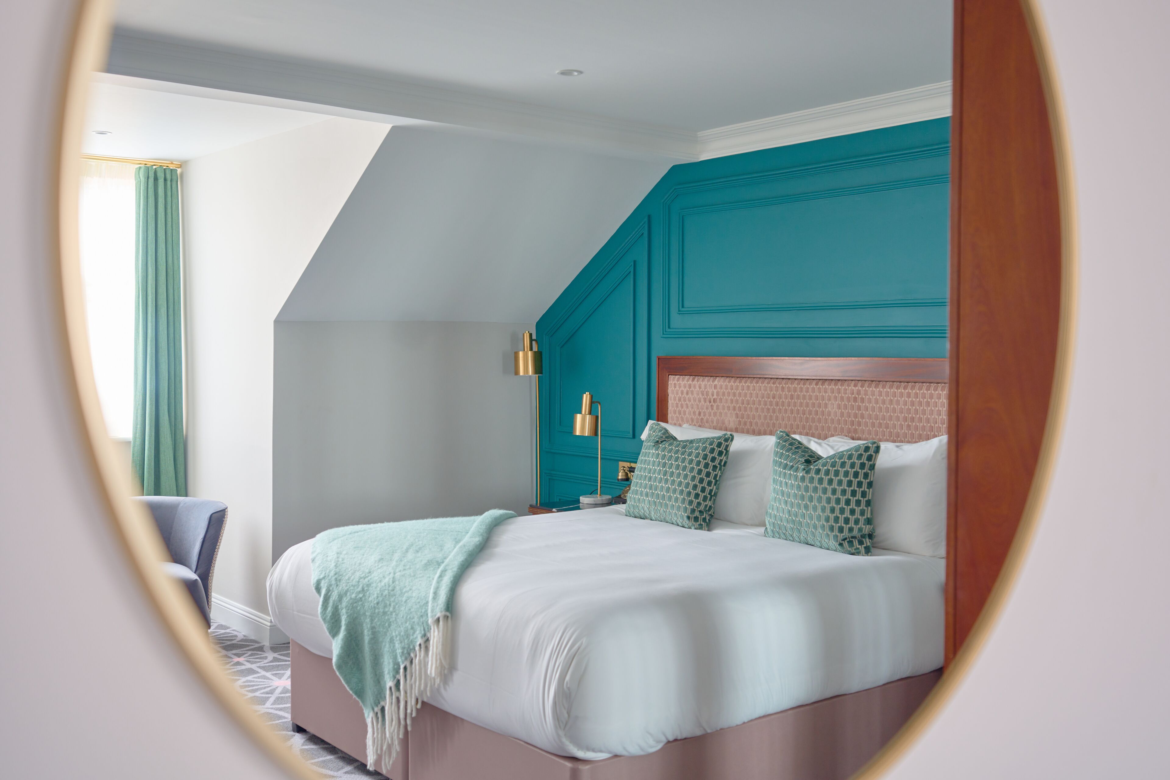 Un dormitorio moderno con techo inclinado, una cama con ropa blanca y almohadas de estampado verde, una pared de acento color turquesa y una mesita de noche pequeña con una lámpara.