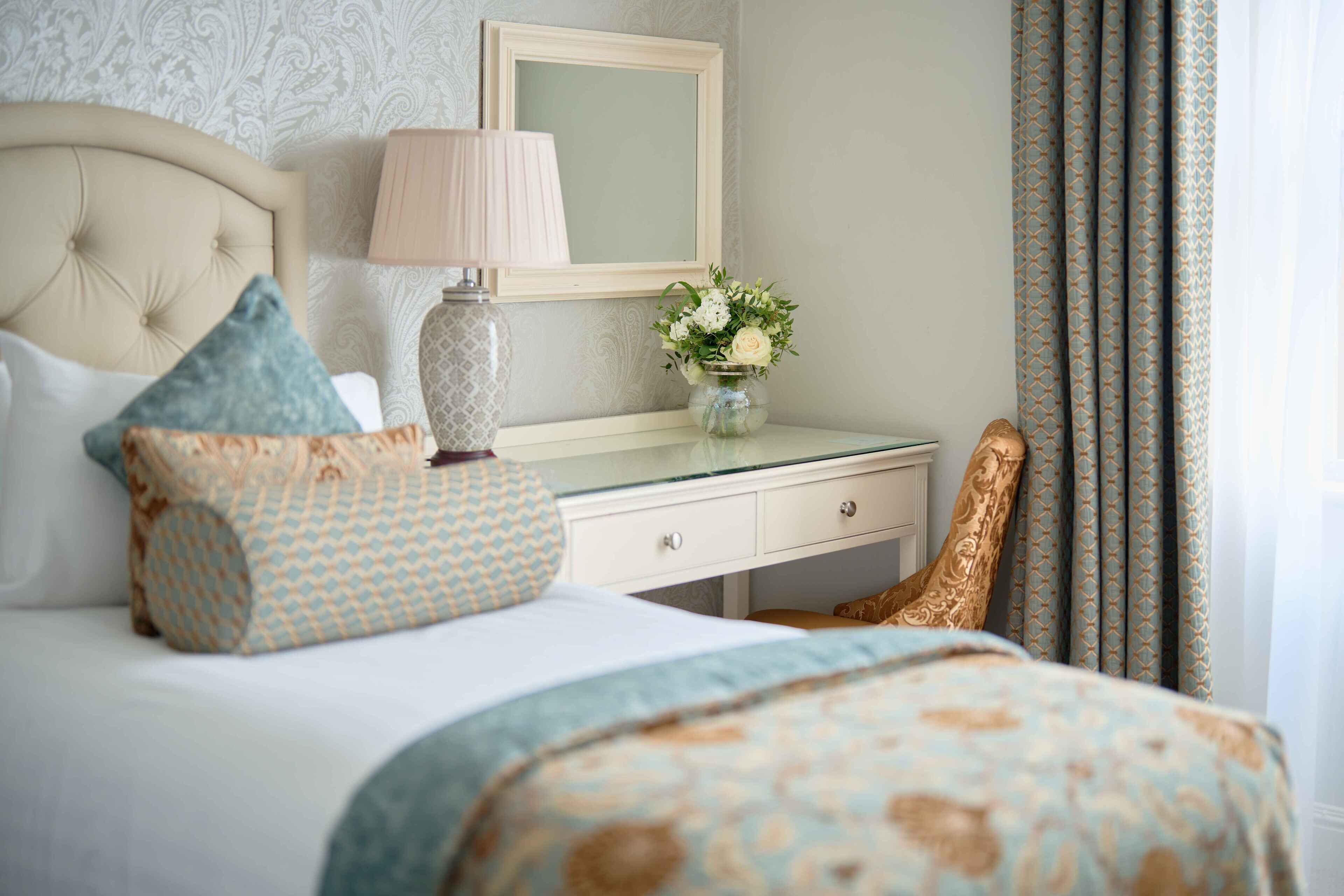 Un dormitorio ordenado con cama, mesita de noche, lámpara, espejo y un jarrón con flores.