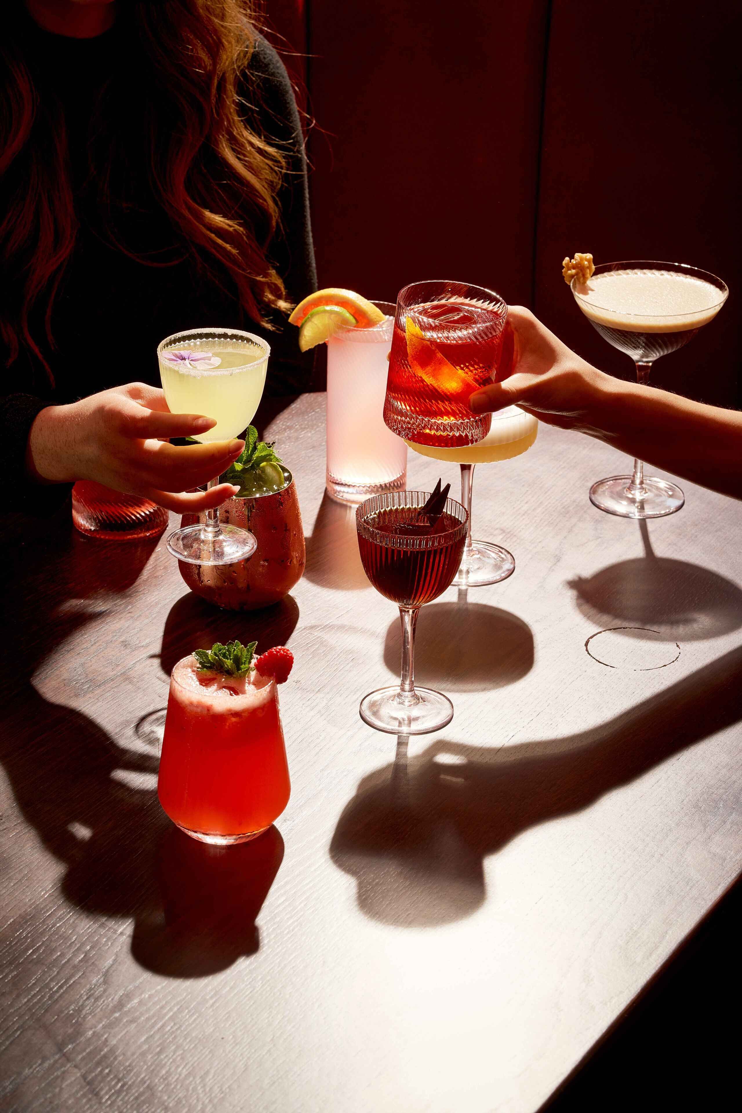 A table with various drinks, including cocktails and a non-alcoholic beverage, with a person holding a glass.