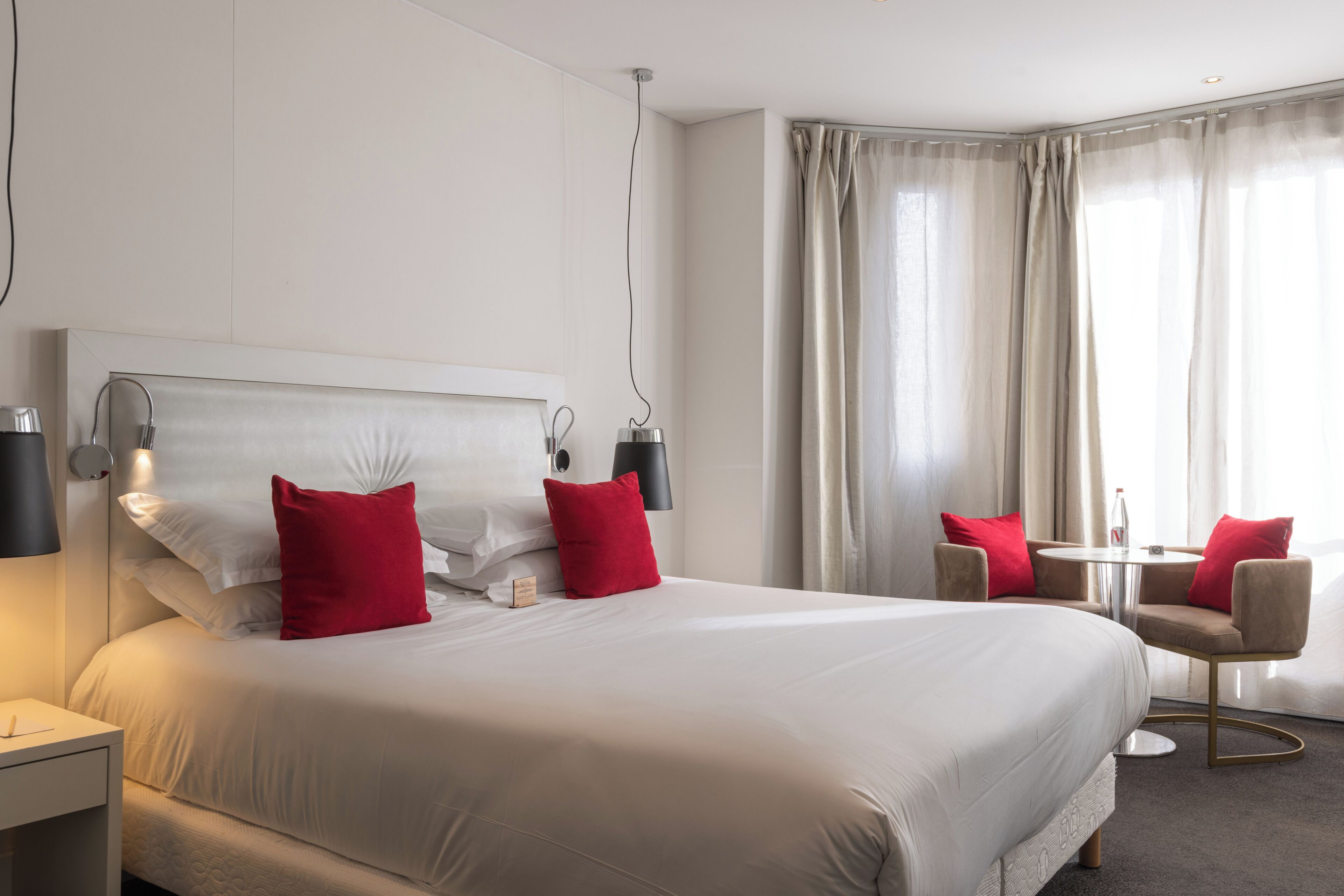Ein Hotelzimmer mit einem großen Bett, zwei roten Kissen, einem Nachttisch mit Lampe, einem kleinen Tisch mit Stühlen und einem Fenster mit Vorhängen.