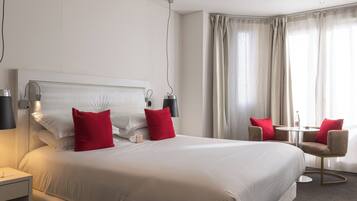 Ein Hotelzimmer mit einem großen Bett, zwei roten Kissen, einem Nachttisch mit Lampe, einem kleinen Tisch mit Stühlen und einem Fenster mit Vorhängen.