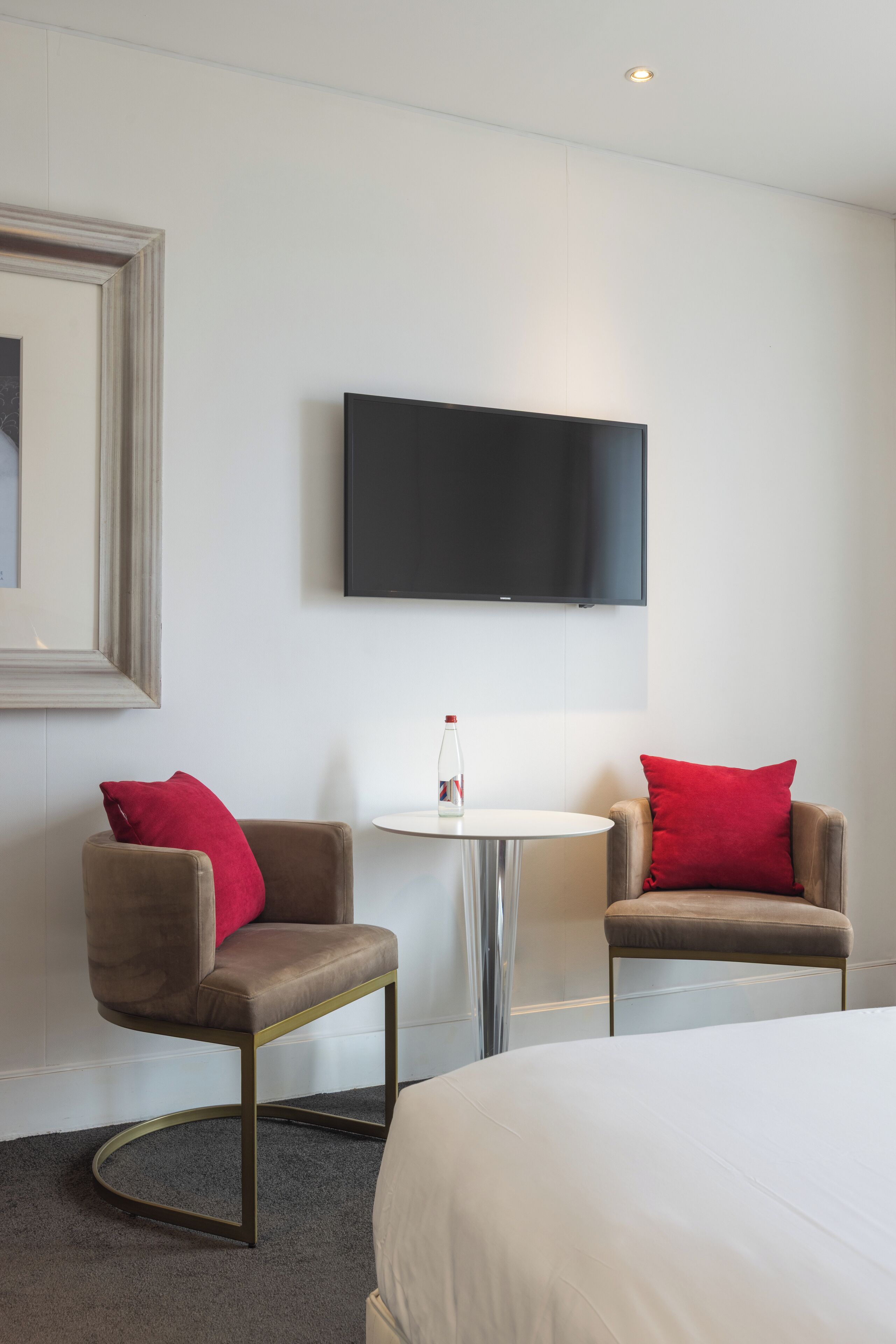 A hotel room with a bed, two armchairs with red cushions, a round table with a bottle, a flat-screen TV mounted on the wall, and a framed picture on the wall.