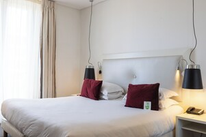 Een hotelkamer met een groot bed, twee bedlampen, een nachtkastje met een telefoon en een raam met gordijnen.