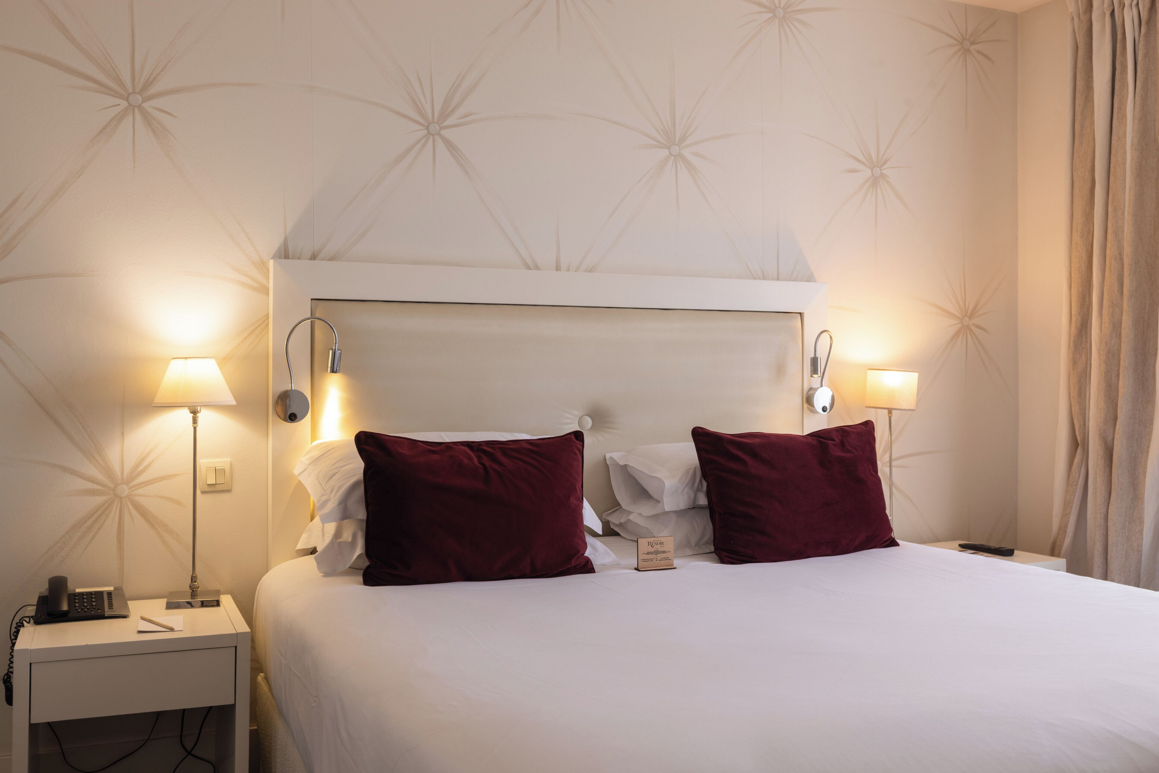 A hotel room with a bed, two red pillows, a nightstand with a lamp, a phone, and a sign indicating the room number.