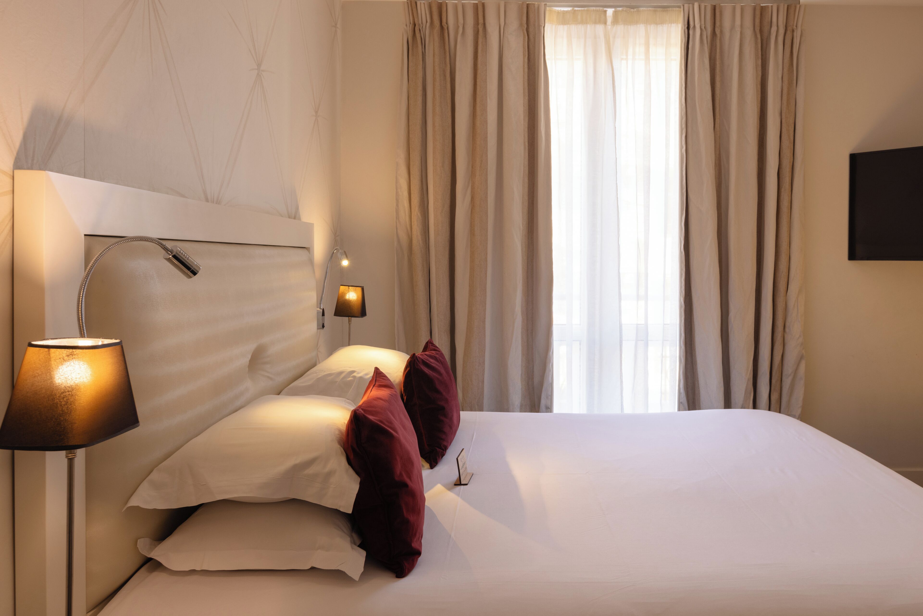 A hotel room with a bed, a lamp, a TV, and a window with curtains.