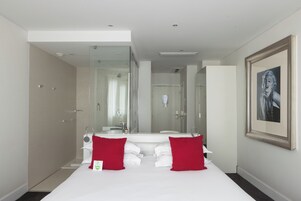 Een moderne hotelkamer met een groot bed, een douchecabine van glas, een grote spiegel en een ingelijst schilderij aan de muur.