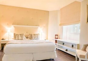 Un dormitorio con una cama grande, mesitas de noche, una mesita auxiliar, una ventana con persianas y una silla blanca.