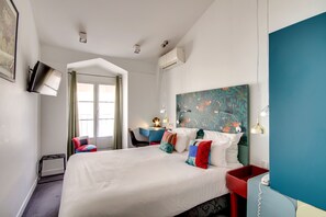 A modern hotel room with a large bed, a desk, and a chair. There is a window with curtains, a painting on the wall, and an air conditioning unit. - Hôtel Sèvres Saint Germain (Paris)